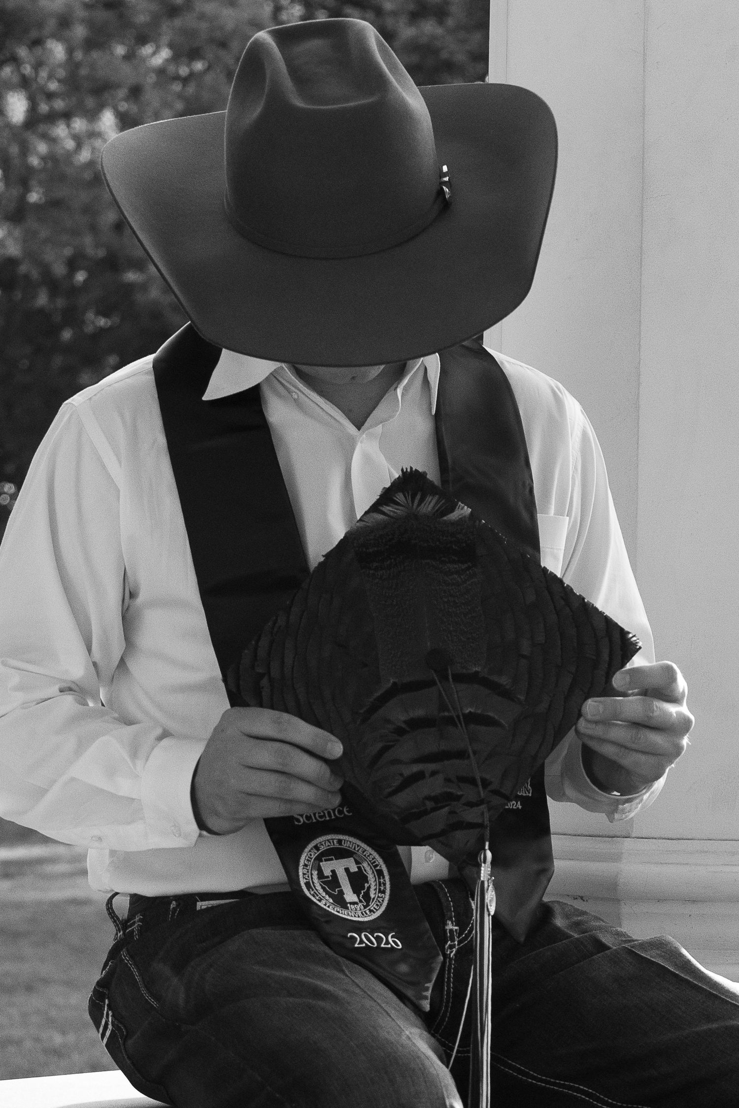 Black and white photo of a cowboy in a white shirt and hat examining a decorative belt buckle while seated outdoors.  STEPHENVILLE TX