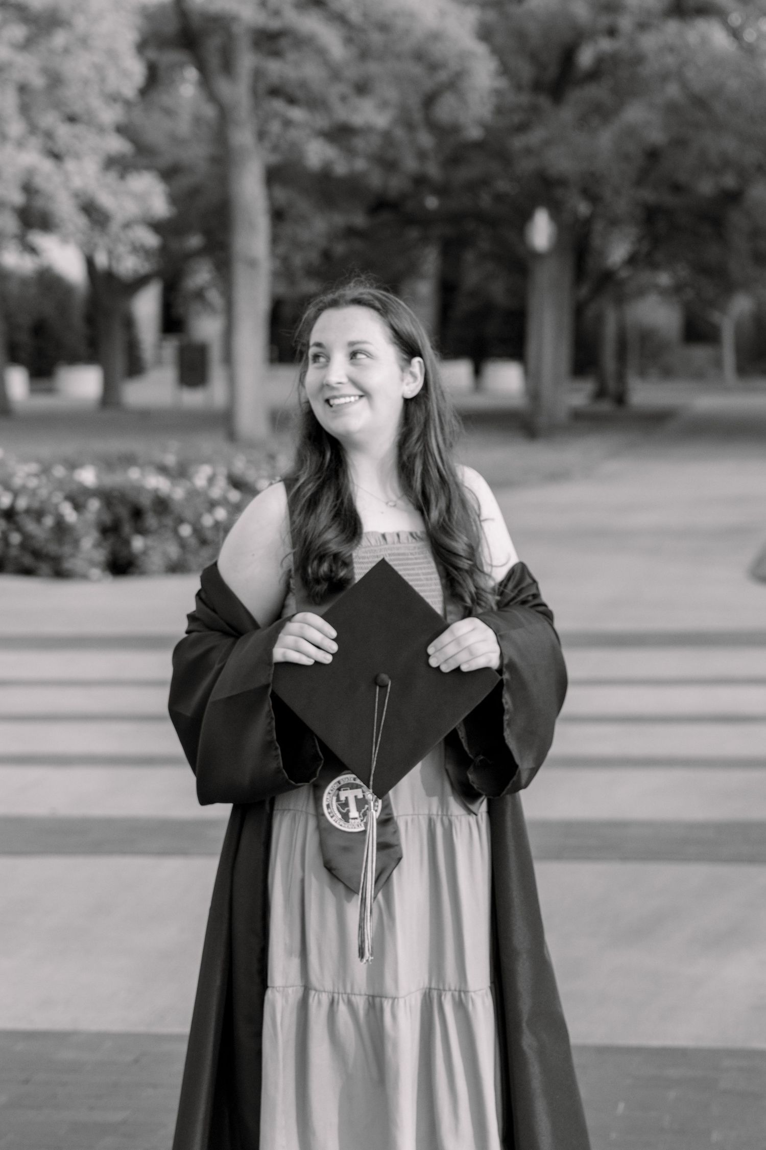 Black and white photo of a smiling graduate in cap and gown holding her graduation cap outdoors. TARLETON STATE UNIVERSITY