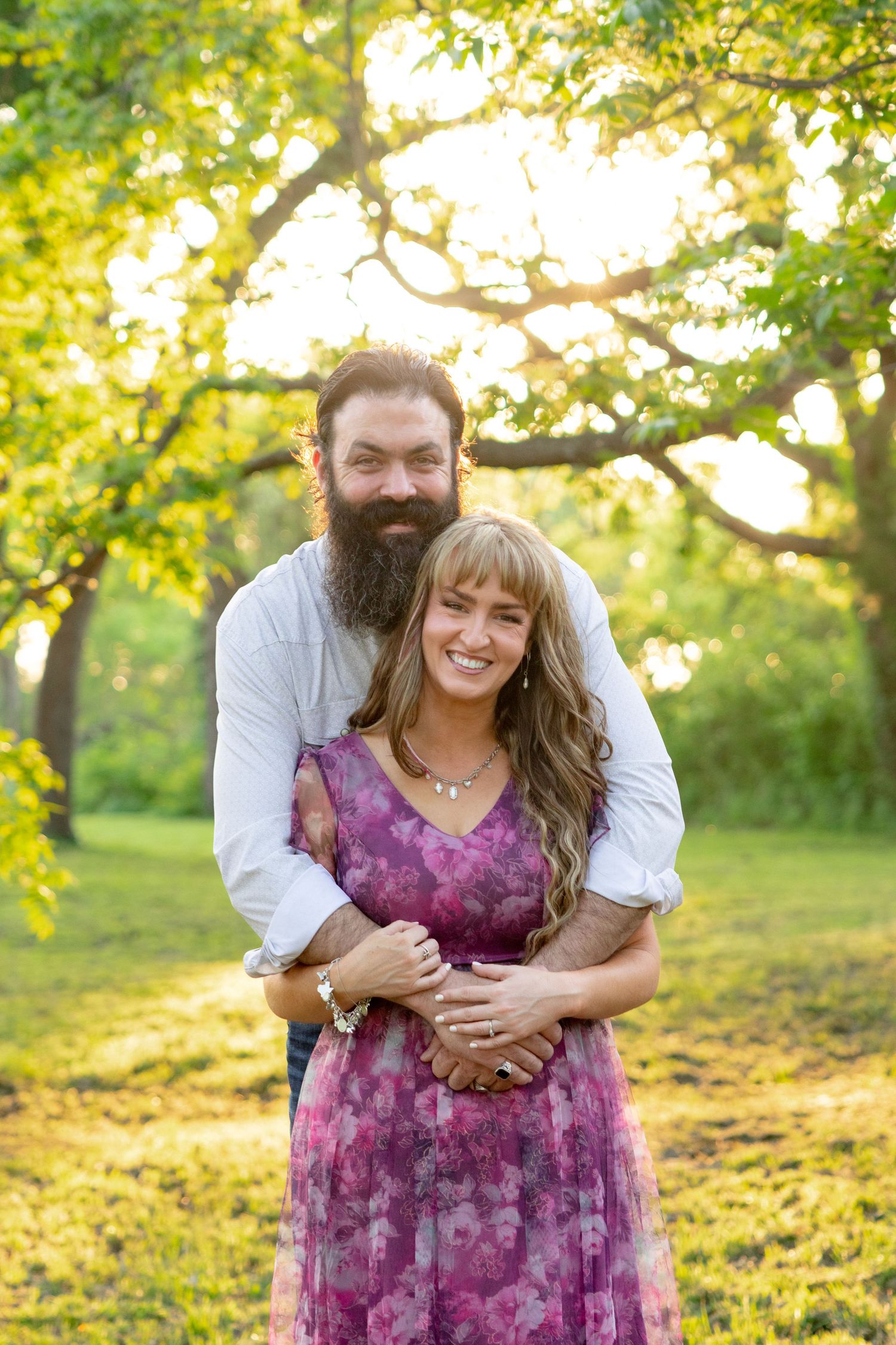 Smiling couple embracing under green trees at sunset, man with beard, woman in purple floral dress. Stephenville Texas