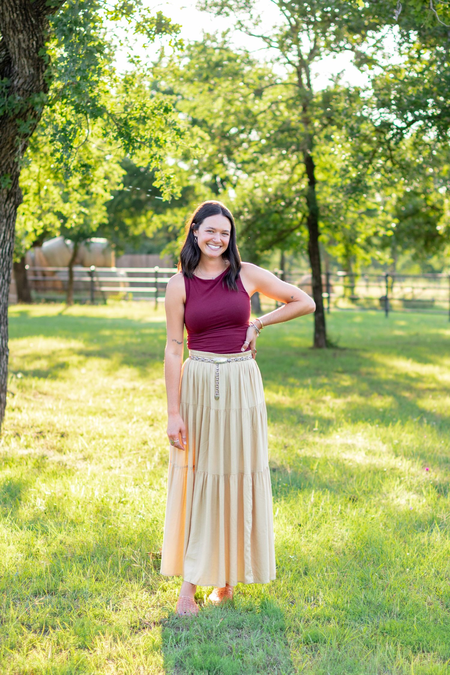 Young woman smiling in burgundy top and gold maxi skirt standing in sunny green park. Stephenville Texas