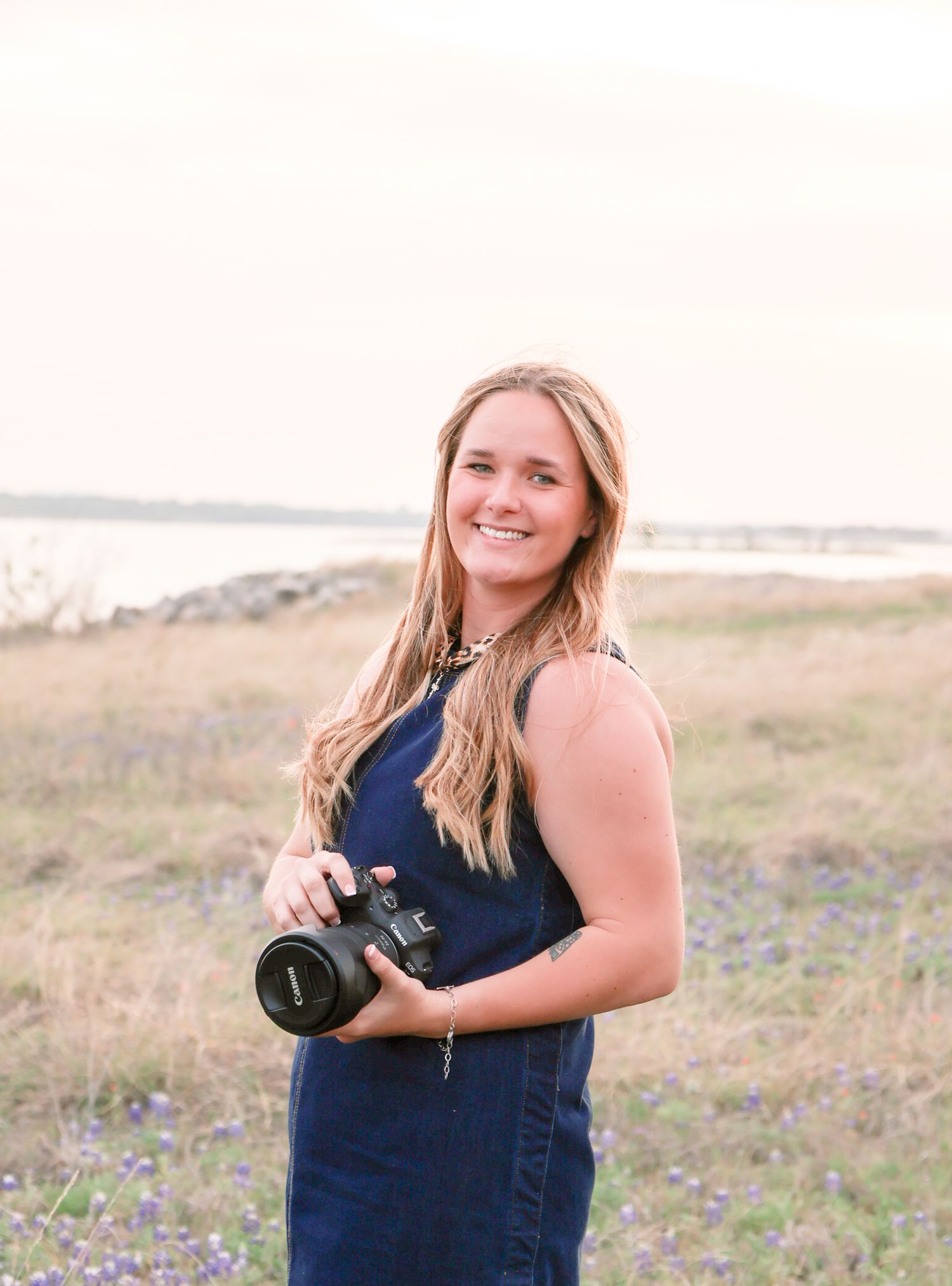 Young female photographer smiles holding Canon camera in Texas bluebonnet field at sunset. Stephenville Texas Photographer
