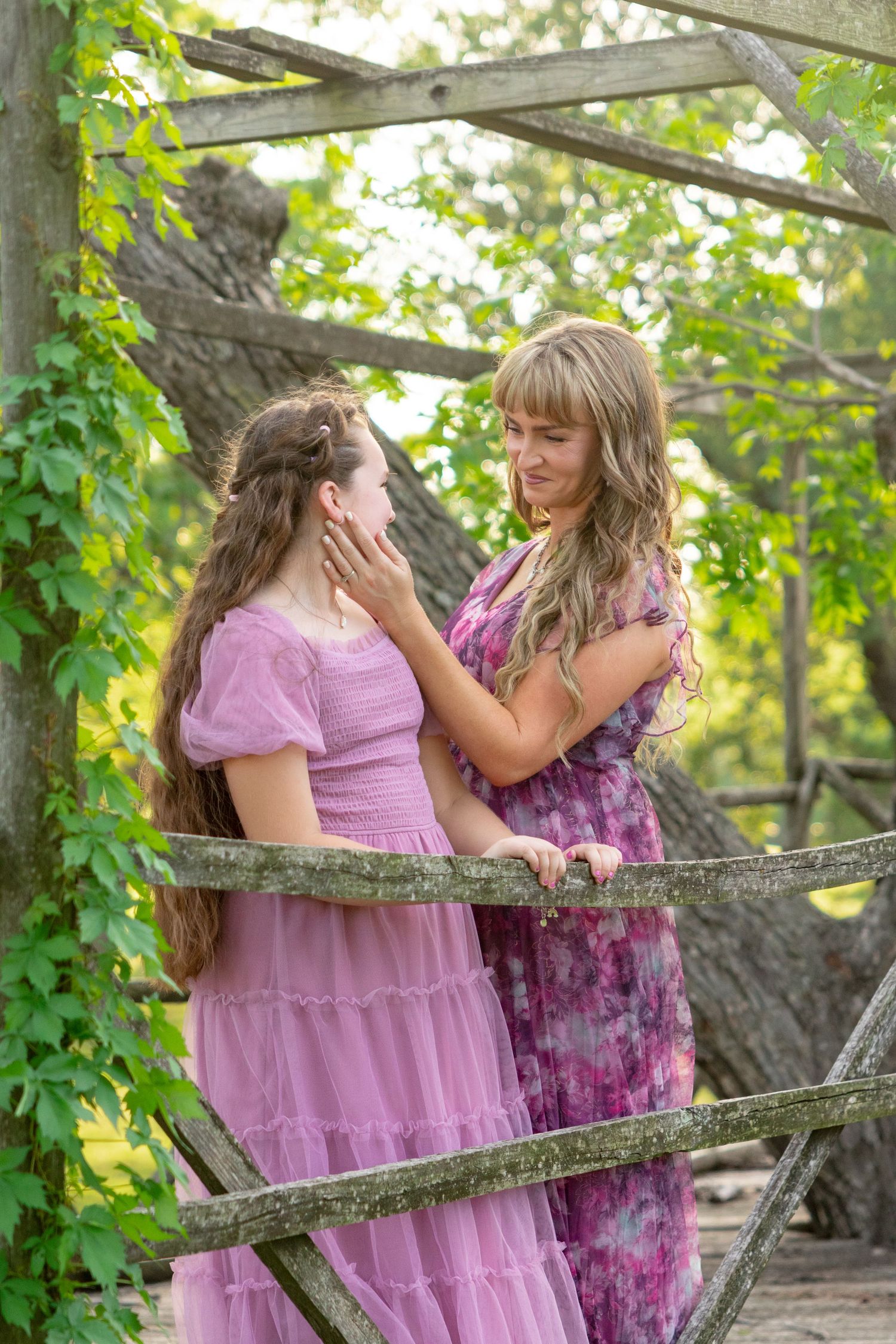 Two women in pink dresses share a tender moment by a rustic wooden fence surrounded by lush green foliage. Stephenville TX Photographer