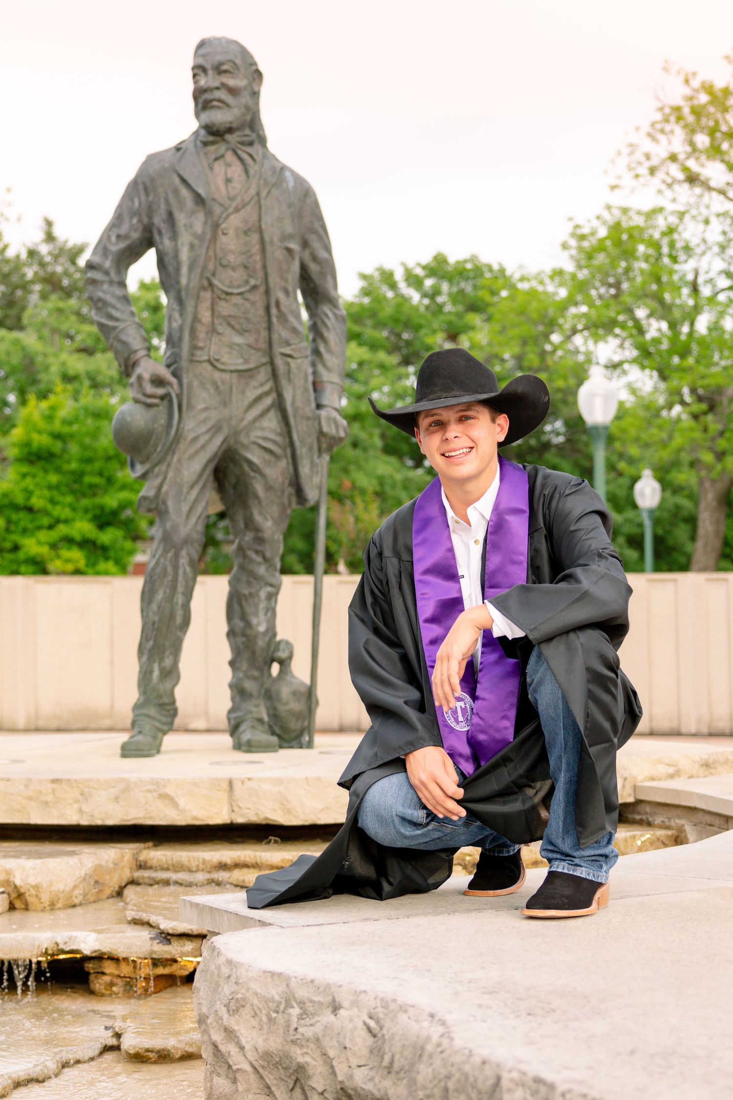 A smiling male graduate in black robes and cowboy hat crouches beside a bronze statue on campus. Stephenville Texas Tarleton state