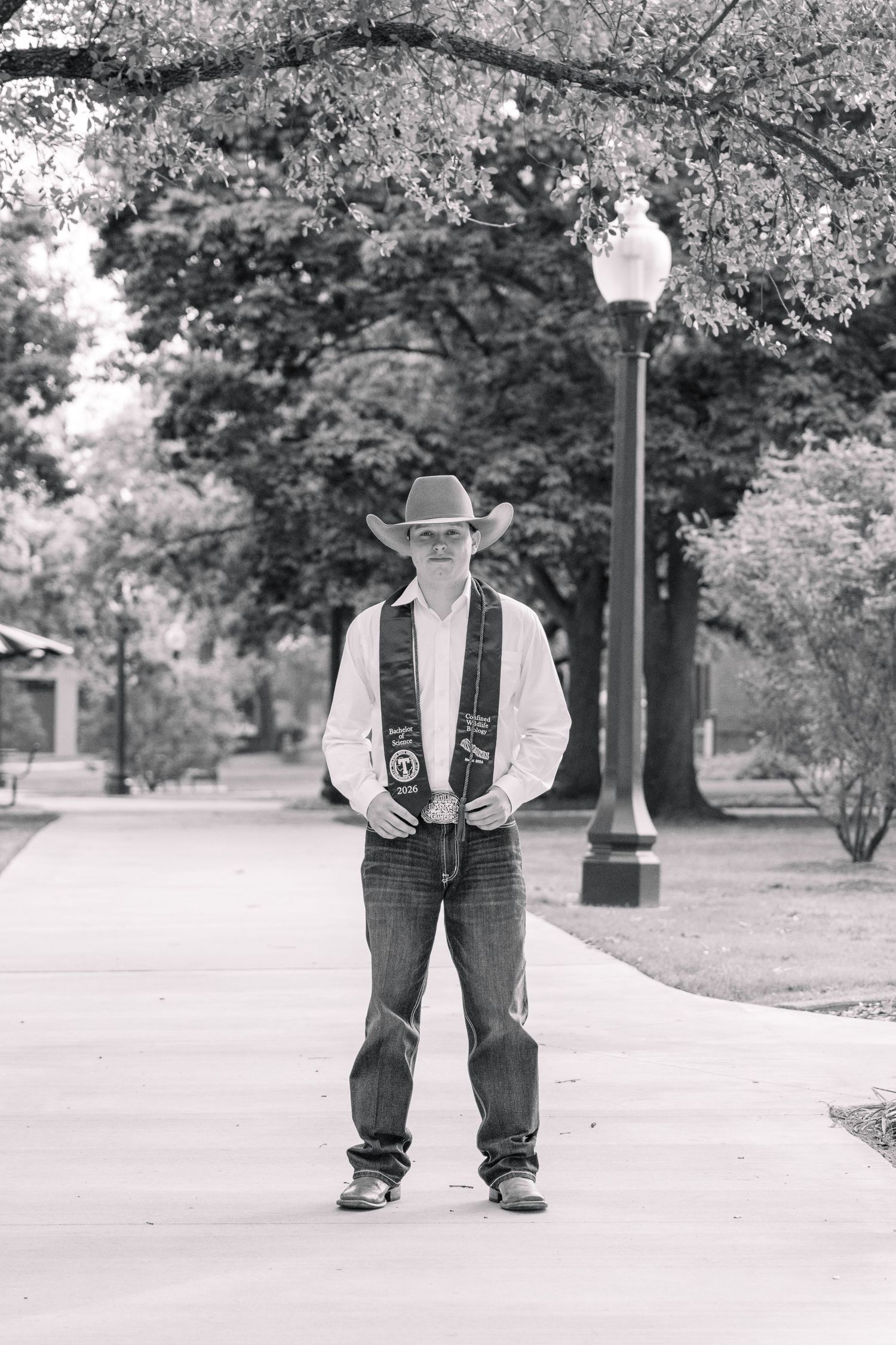 Man in cowboy hat and graduation sash stands on campus walkway in black and white photo. Stephenville Texas