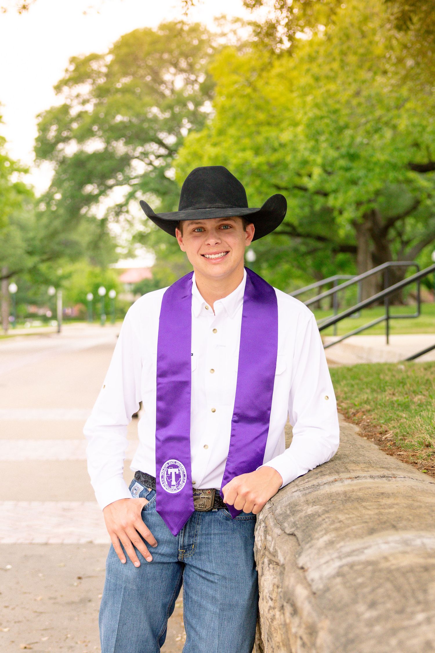 Young man in black cowboy hat and purple graduation stole leans against stone wall outdoors. Stephenville Texas