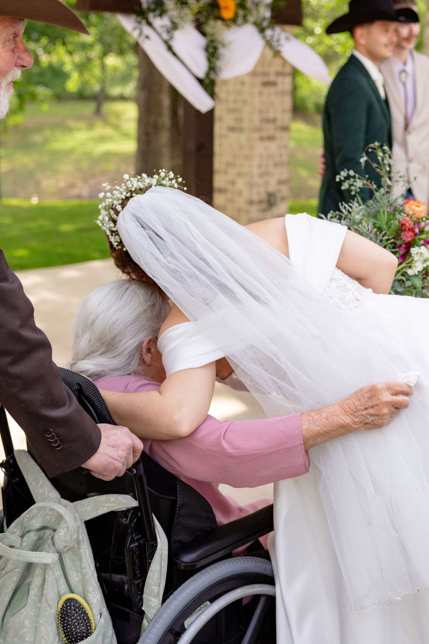 Bride in white gown and veil leans down to embrace elderly woman in wheelchair during wedding ceremony. Granbury Texas