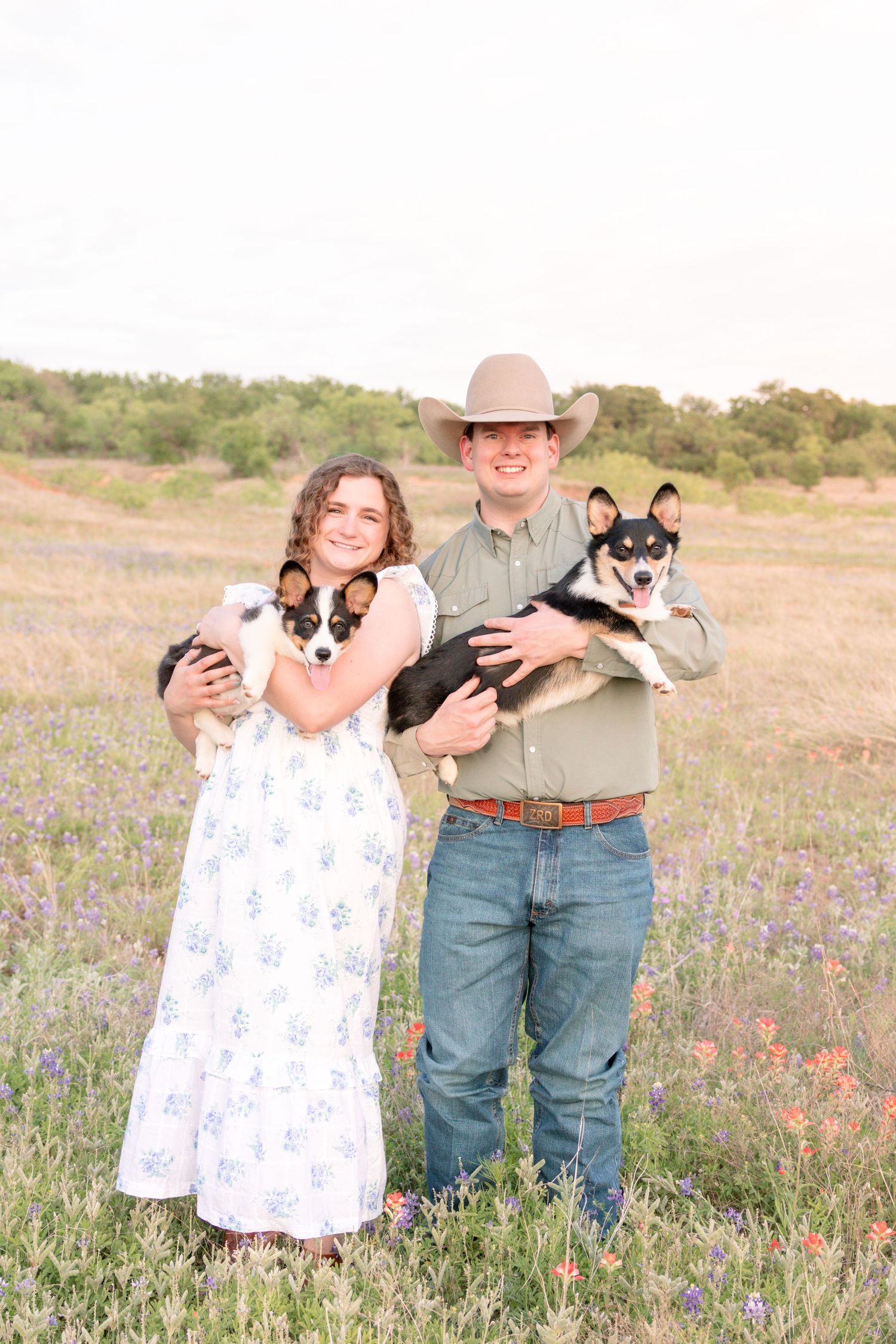 Smiling engaged couple stands in wildflower field holding two Corgi dogs, man wearing cowboy hat and woman in white dress. Stephenville Texa