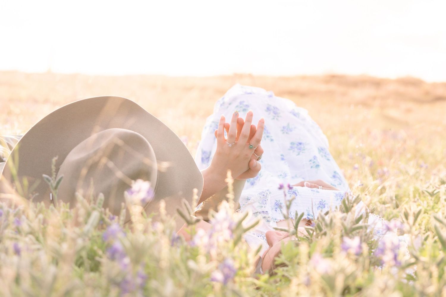 Person in floral dress sits among purple wildflowers in sunlit field beside a wide-brim hat. Stephenville