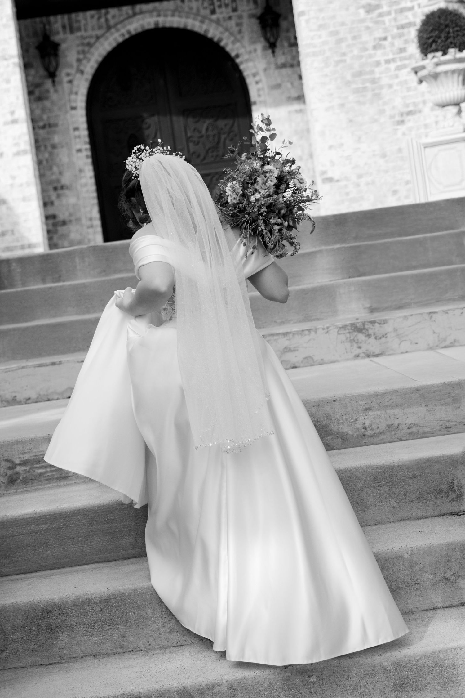 Black and white photo of bride in white dress and veil ascending stone church steps. Granbury Texas