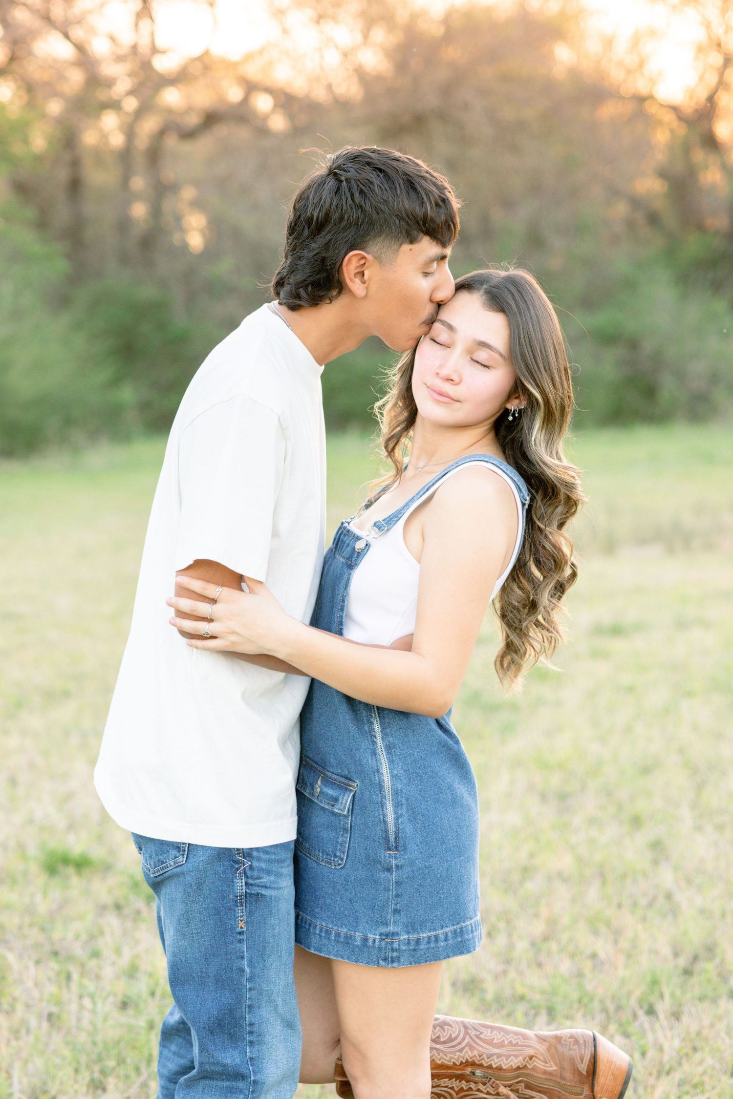 Young couple in a field at sunset, boy kissing girl on forehead, girl wearing denim overalls and cowboy boots.