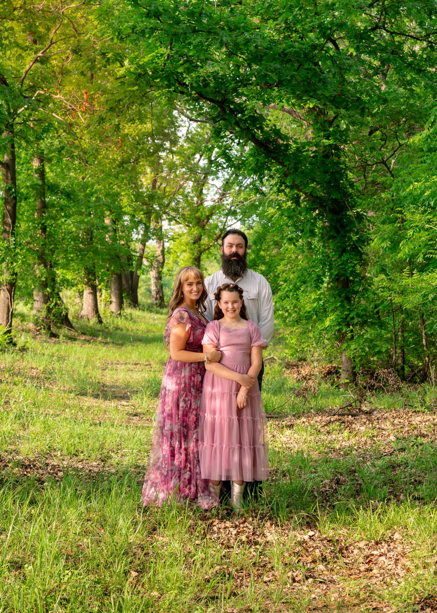 A bearded father, mother, and young daughter pose together in matching pink outfits in a lush green forest path.