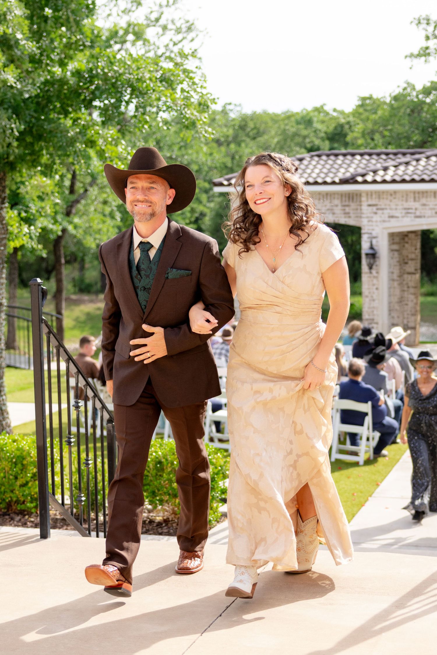 Smiling couple in western attire walk arm-in-arm down outdoor wedding venue path after ceremony. Granbury Texas