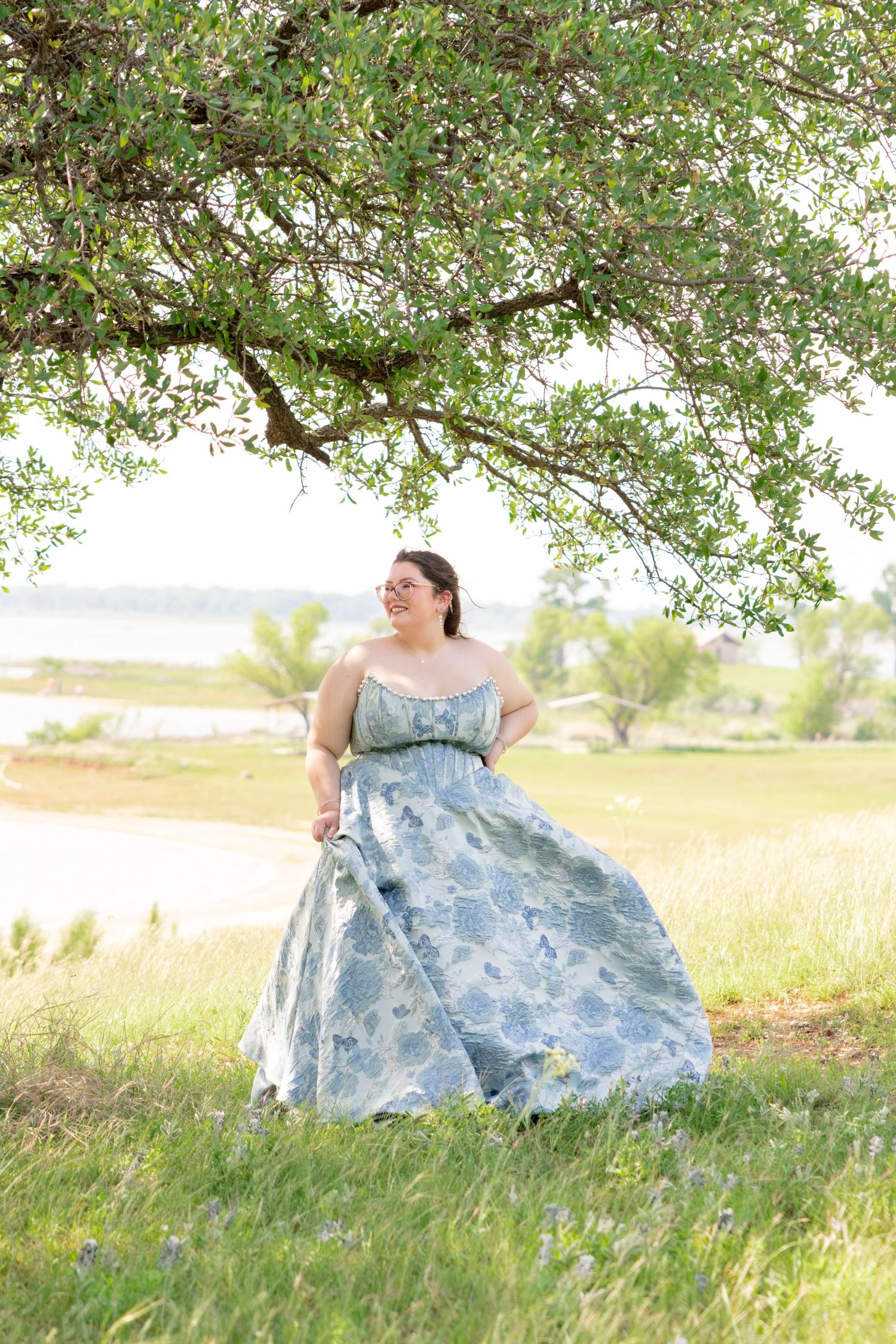A young woman in a blue floral strapless ballgown stands under a blooming tree near a peaceful lakeside. Stephenville Texas