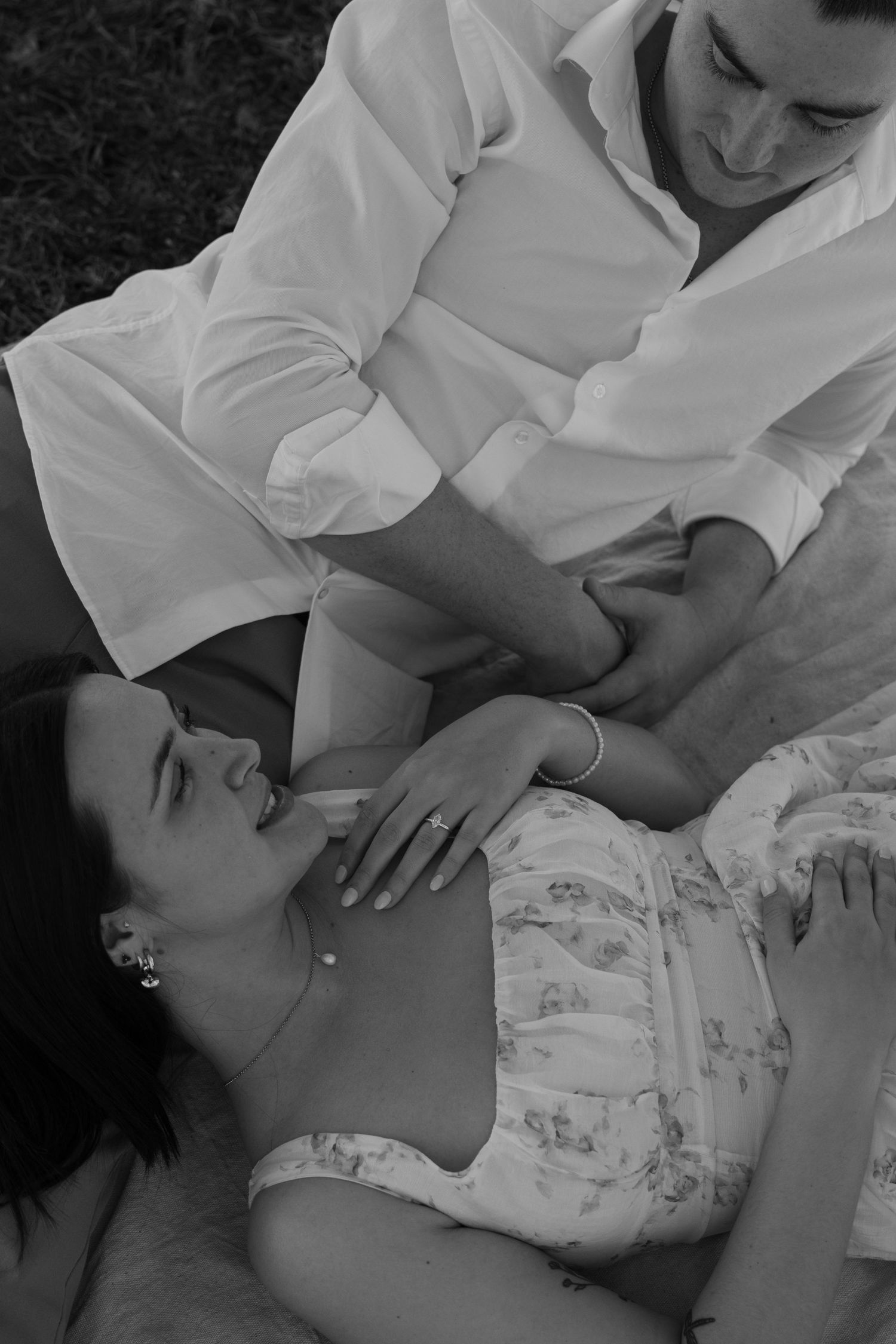 Black and white romantic photo of couple relaxing in grass, woman lying back laughing as man in white shirt gazes at her. Fredericksburg
