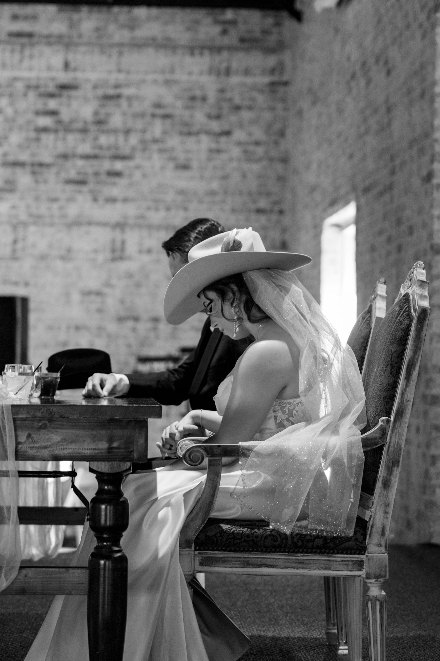 Black and white photo of bride in wide-brim hat and veil sitting at table with groom at wedding reception. Granbury Texas