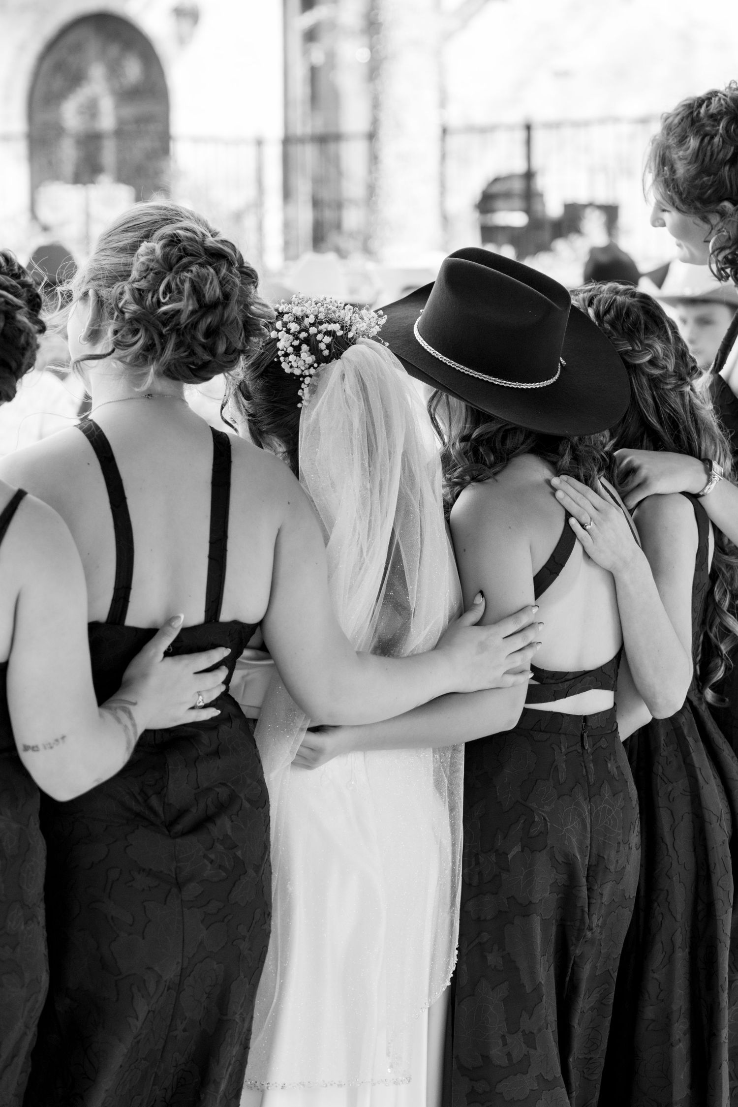 Black and white photo of bride and wedding party in hats huddled together in group embrace outdoors. Granbury Texas