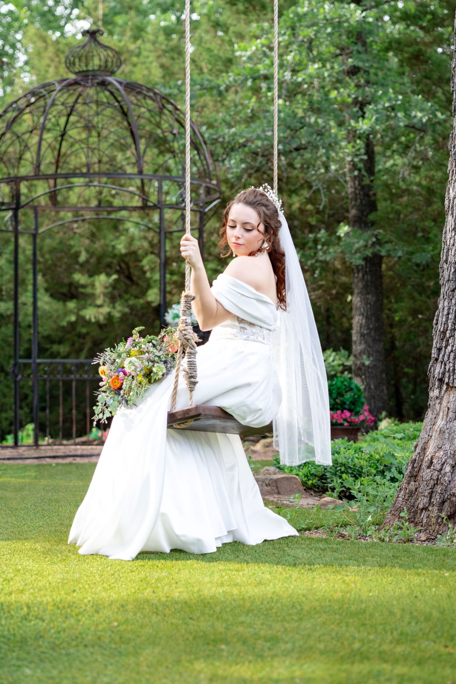 Bride in off-shoulder wedding gown sitting on a rope swing holding a colorful bouquet.