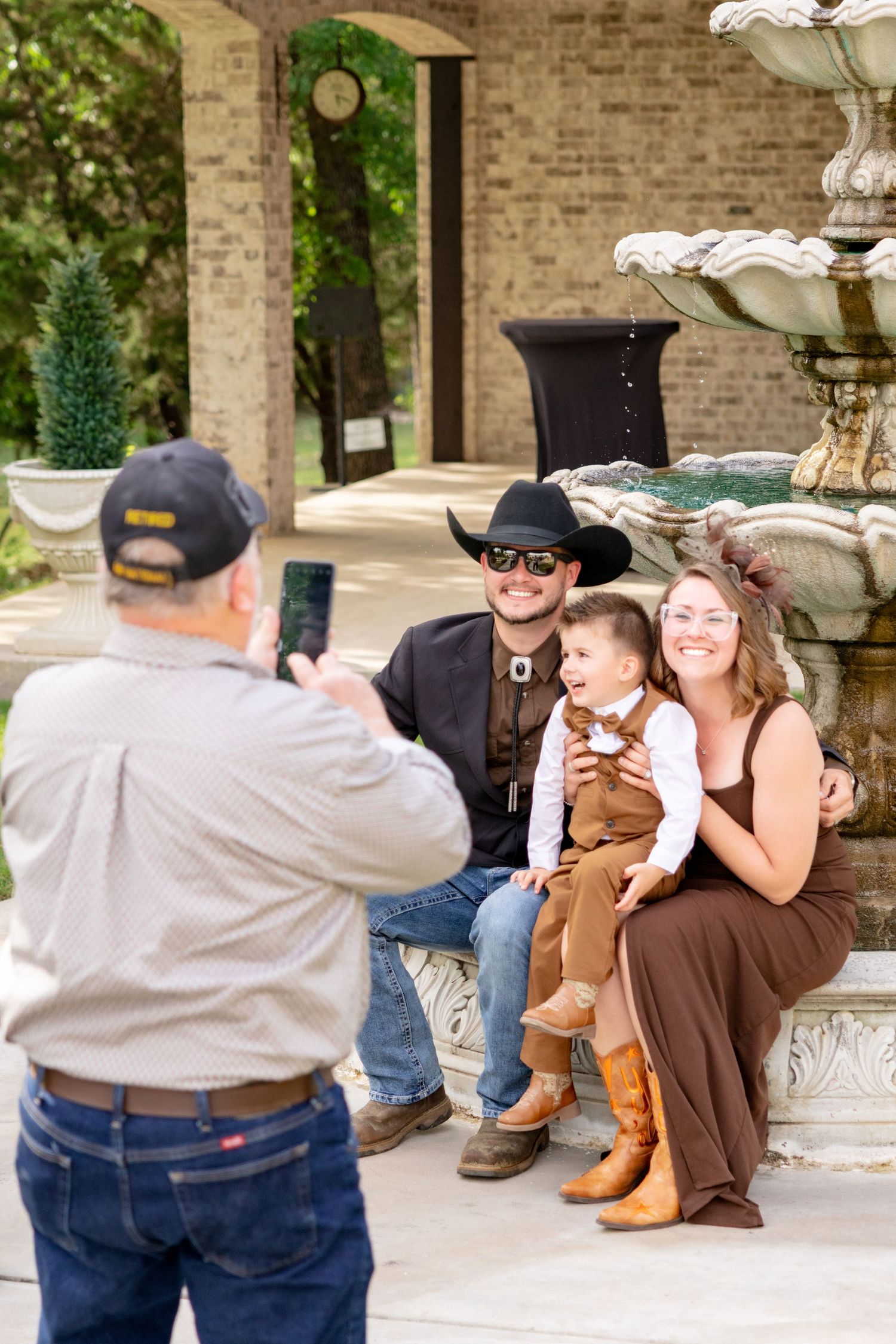 Older man photographs smiling couple in cowboy hats with young child by stone fountain at wedding. Granbury Texas