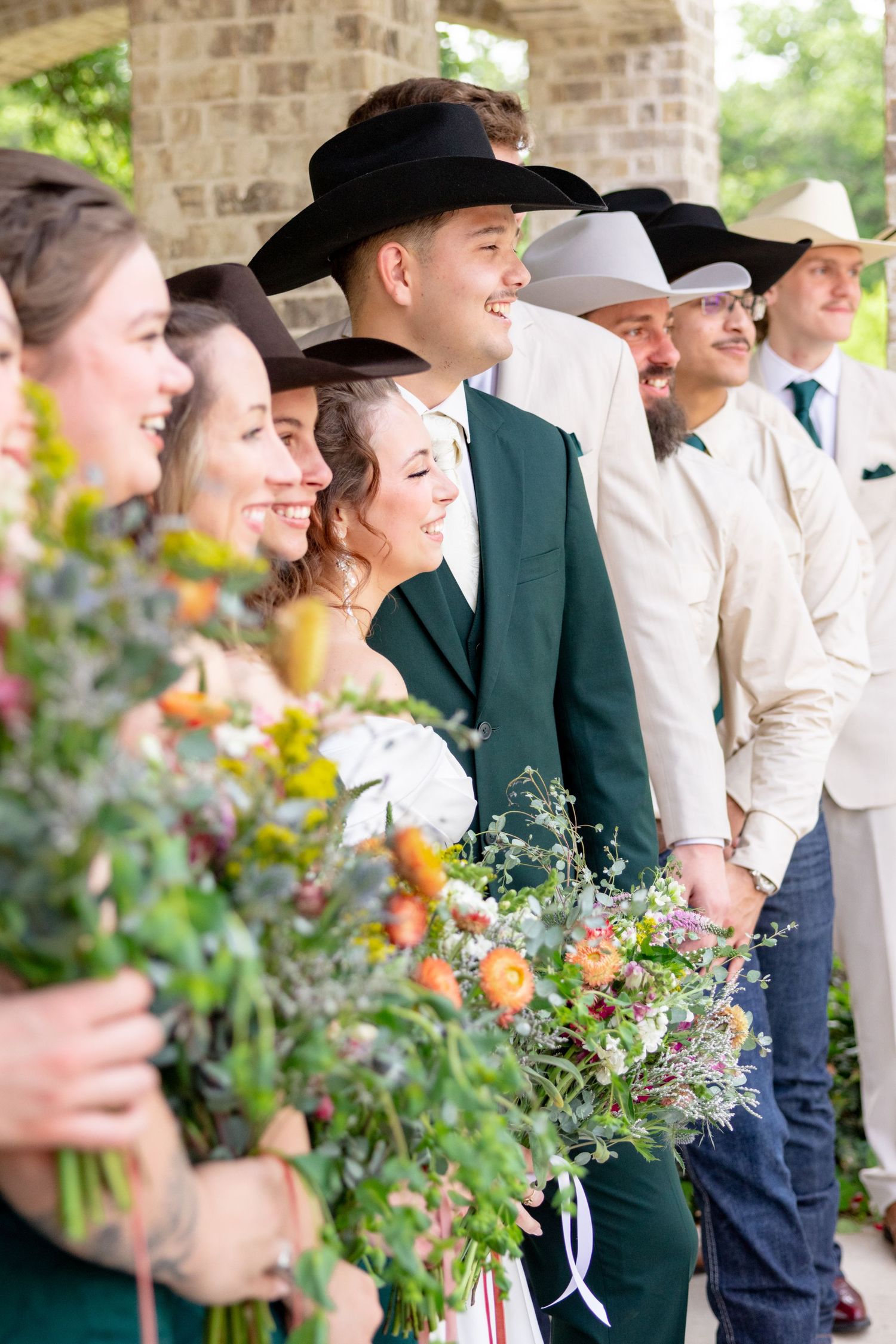 Wedding party in cowboy hats holding colorful wildflower bouquets lined up under stone archway. Granbury Texas
