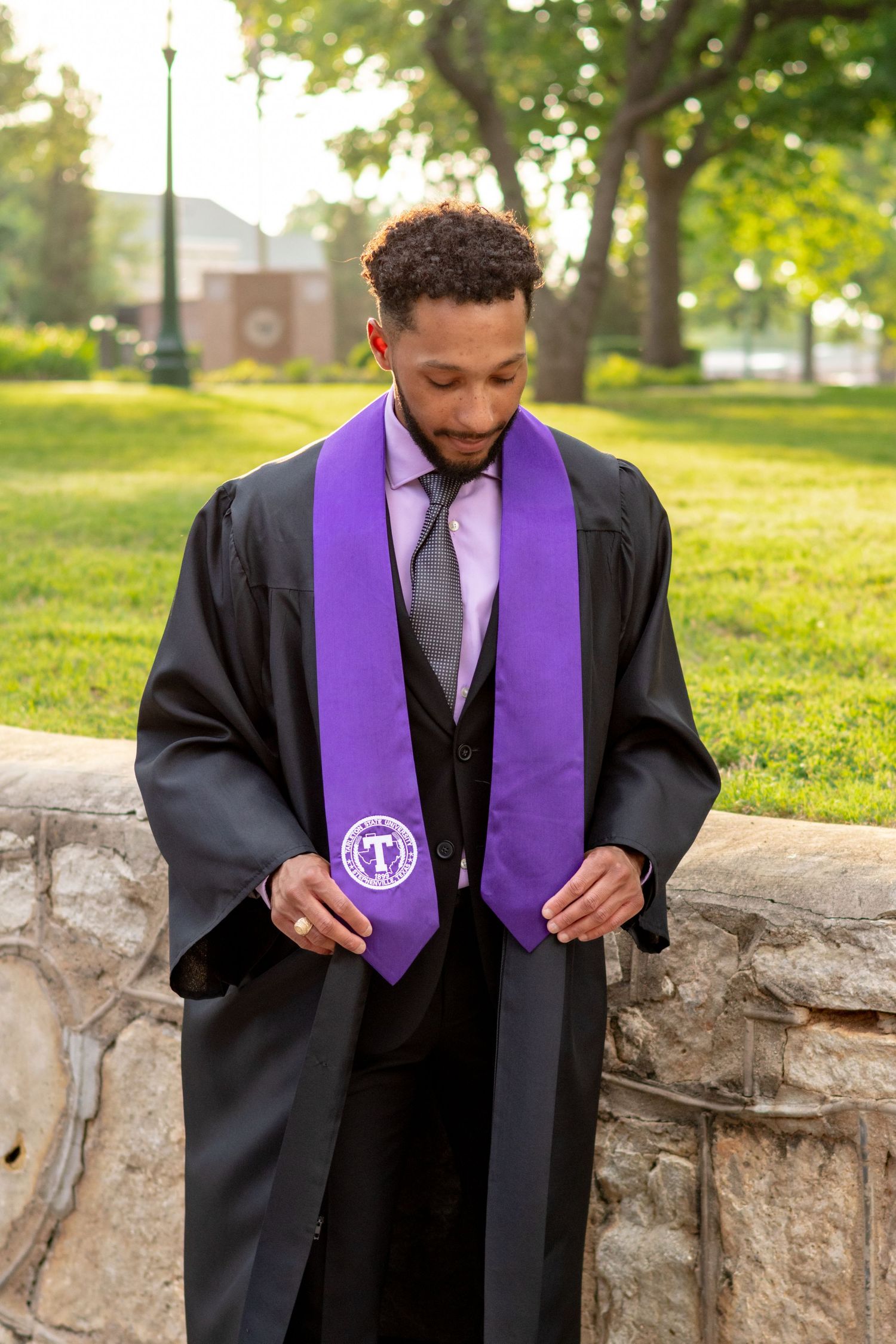 Young man in black graduation gown with purple stole posing outdoors on a sunny day. TARLETON STATE UNIVERSITY