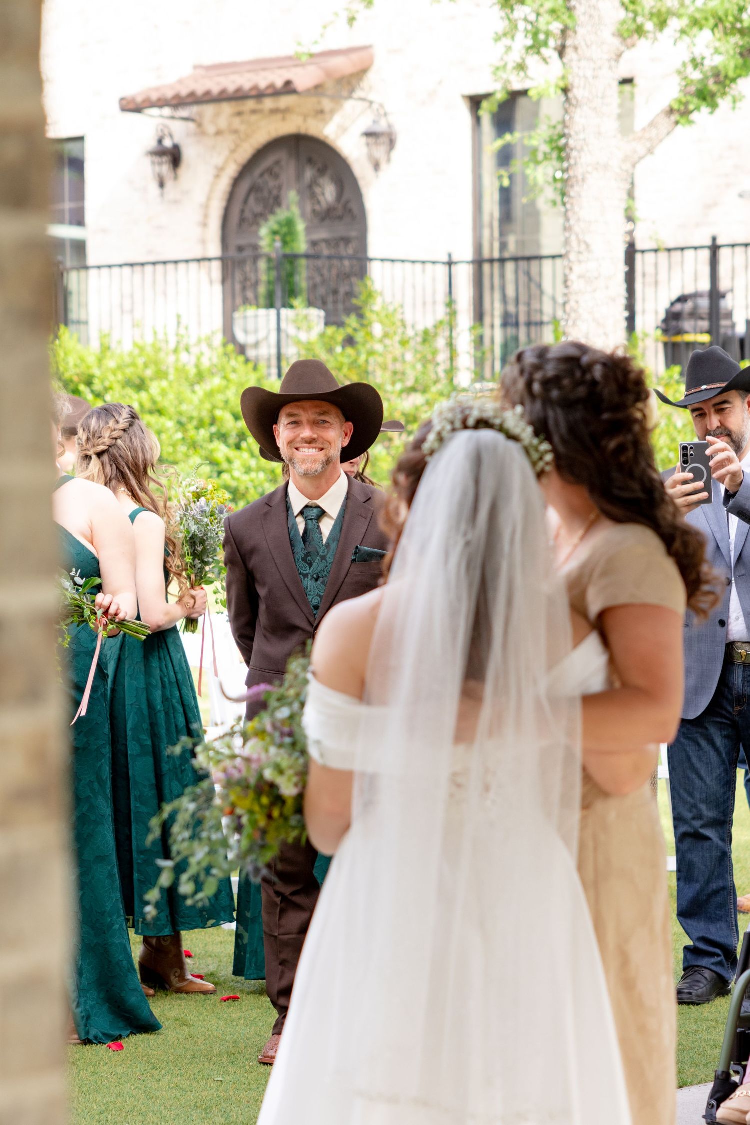 Groom in brown suit and cowboy hat smiles warmly as bride in white veil walks toward him at altar. Granbury Texas