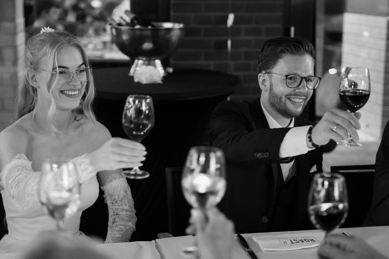 Black and white photo of a smiling bride and groom raising wine glasses during a wedding reception toast.