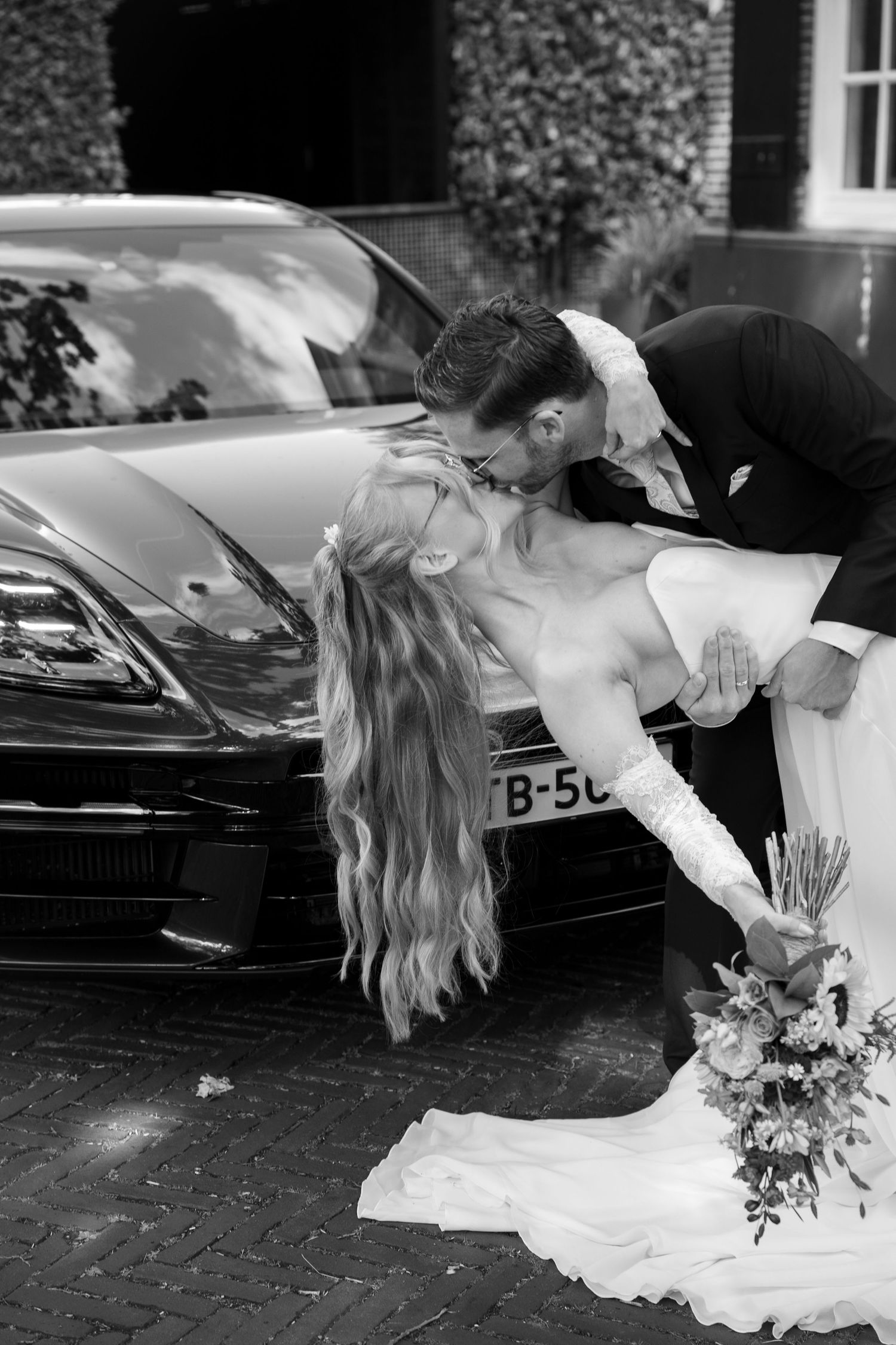 Black and white photo of a bride and groom sharing a romantic dip kiss in front of a luxury car.