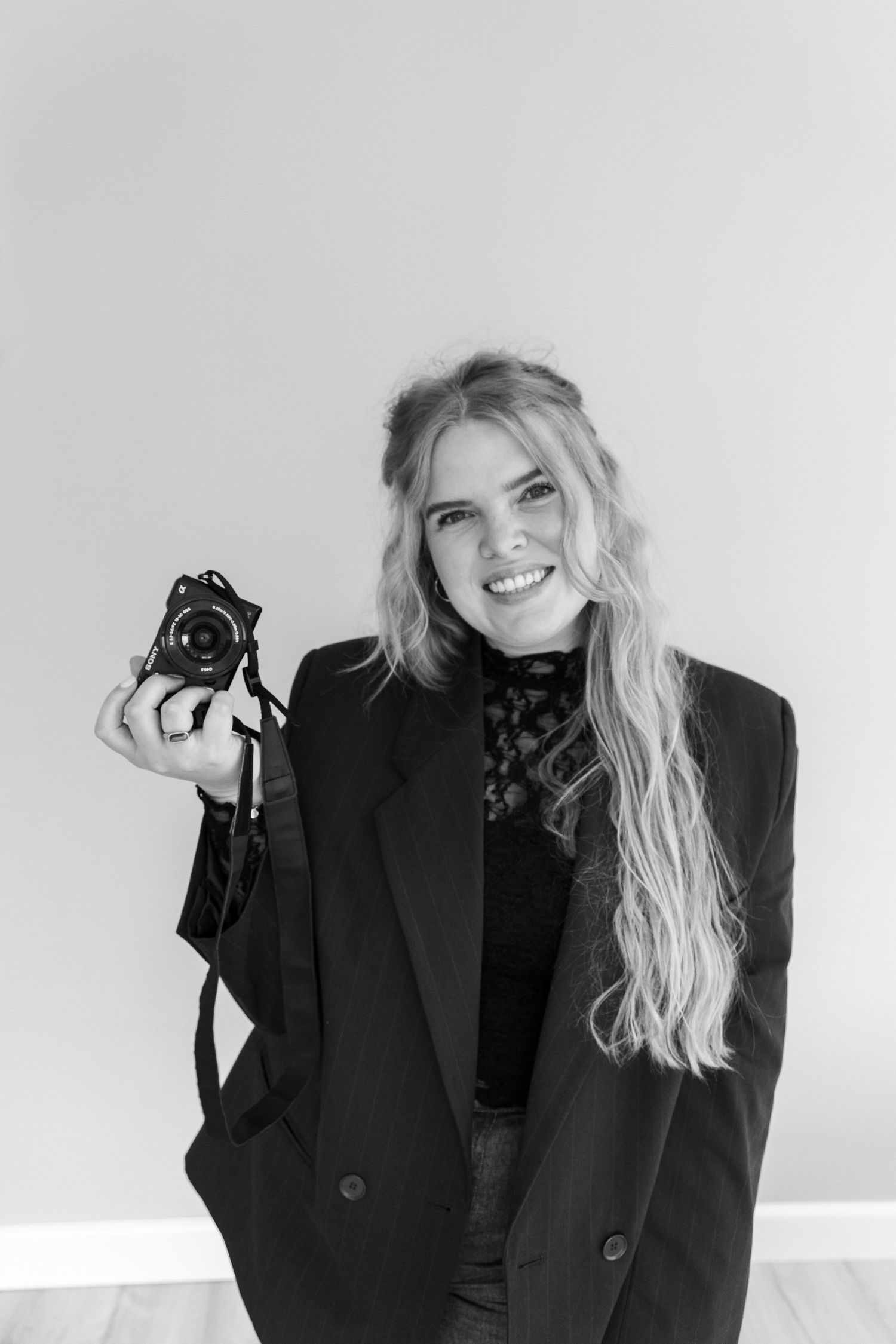Black and white photo of smiling blonde woman holding camera in black blazer.