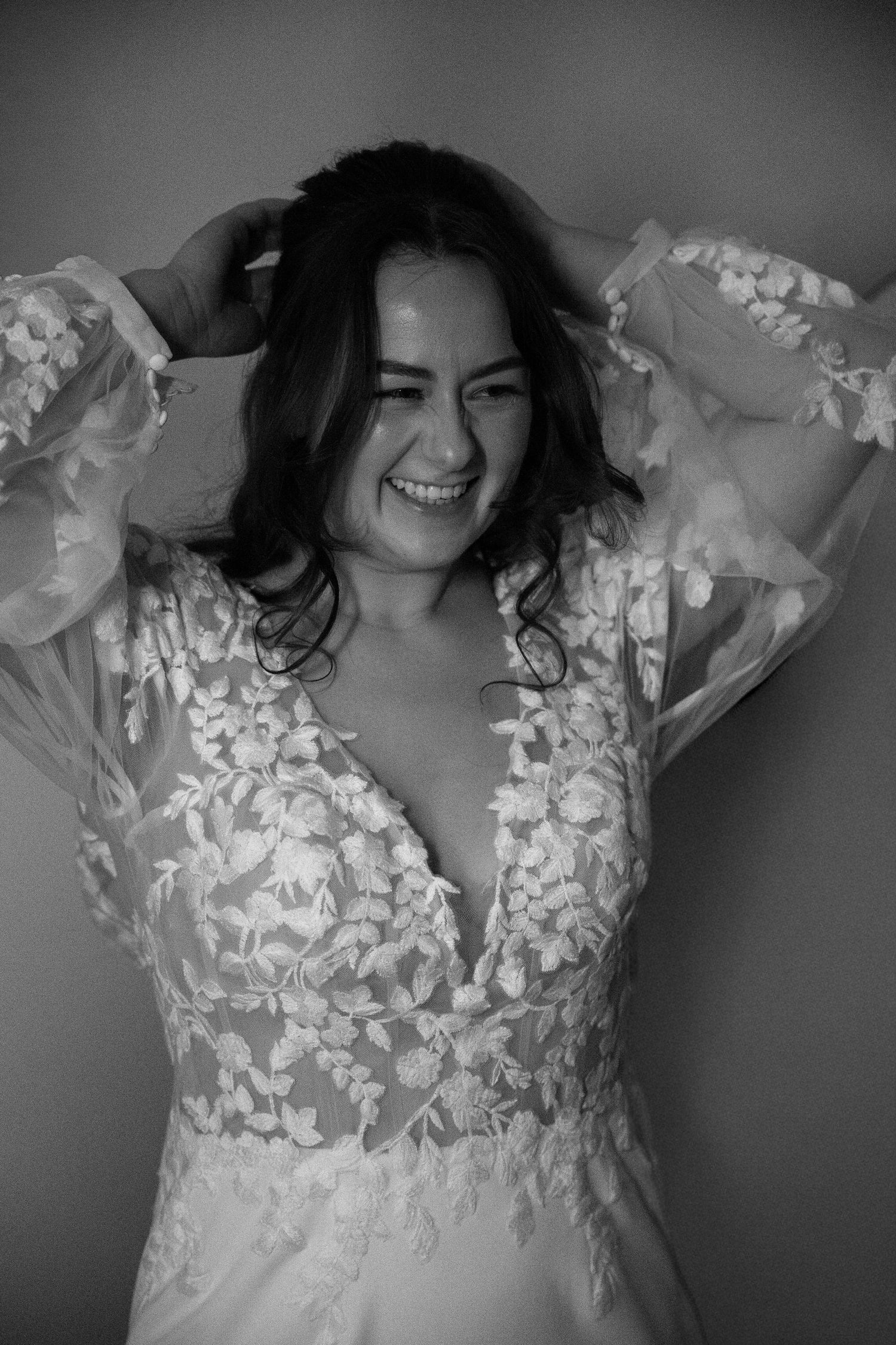 Smiling bride in floral lace wedding gown with sheer sleeves, hands in hair, black and white portrait photo.
Sjoerd en Sanne - Bruiloft