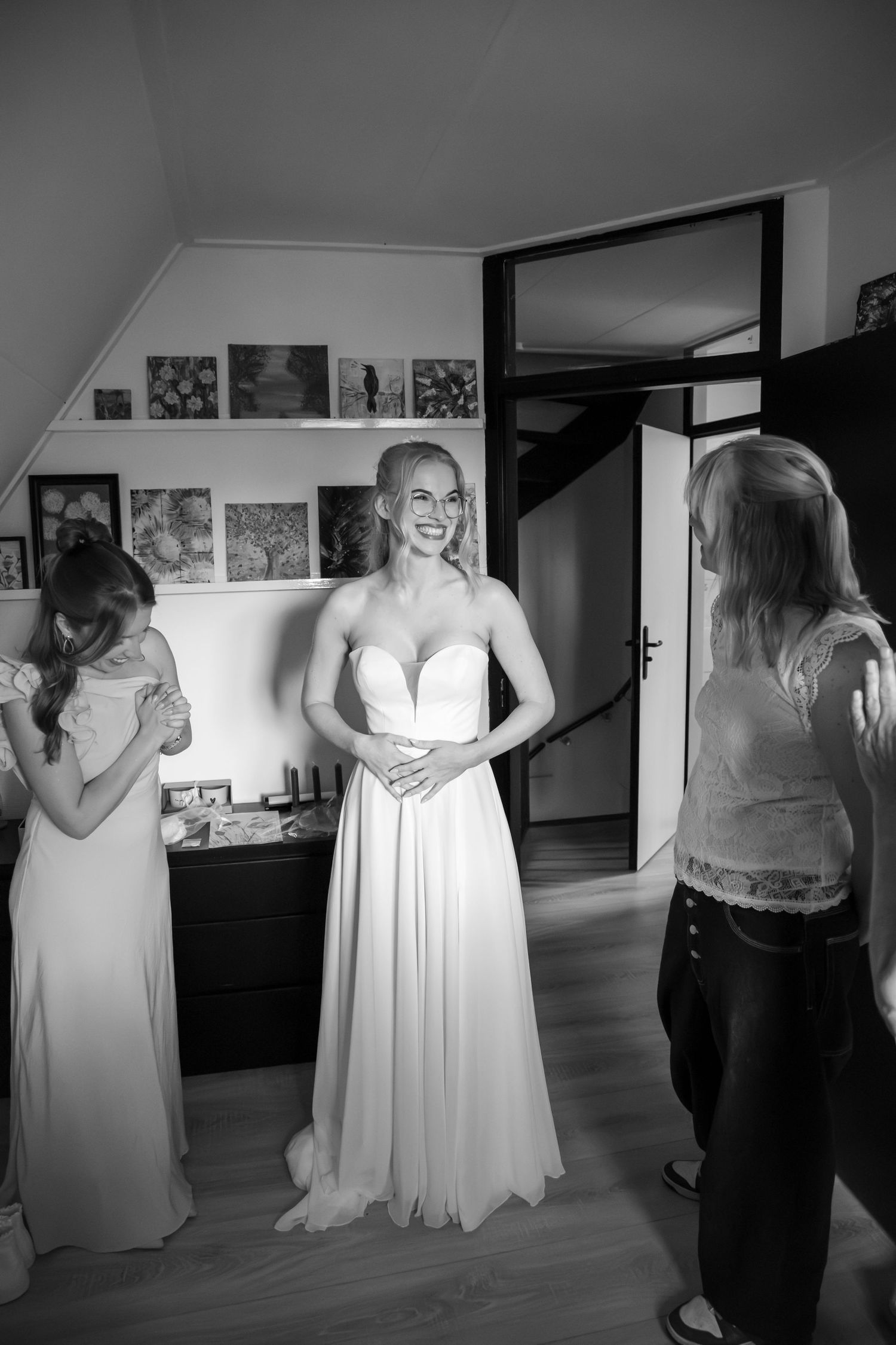 Smiling bride in white strapless gown stands with sister and friend during wedding preparations in black and white photo.