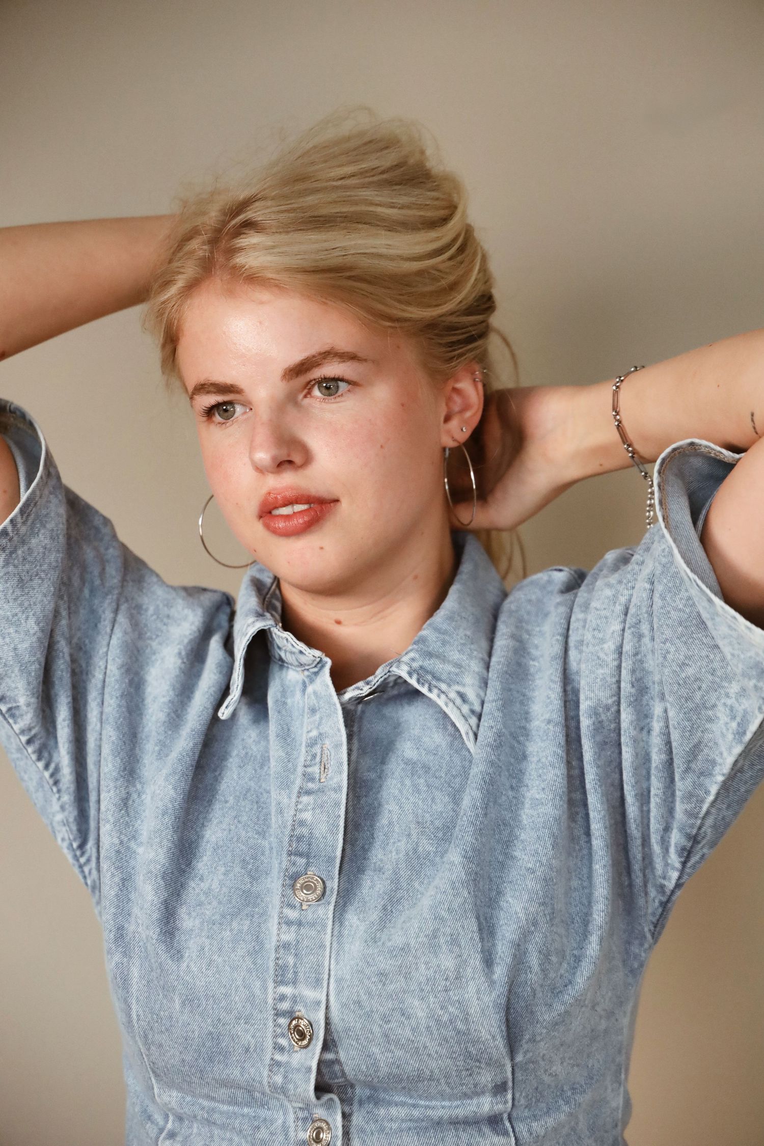 Young blonde woman in acid wash denim shirt with hoop earrings and bracelet, hands behind head, neutral background.
Tess - Soloshoot