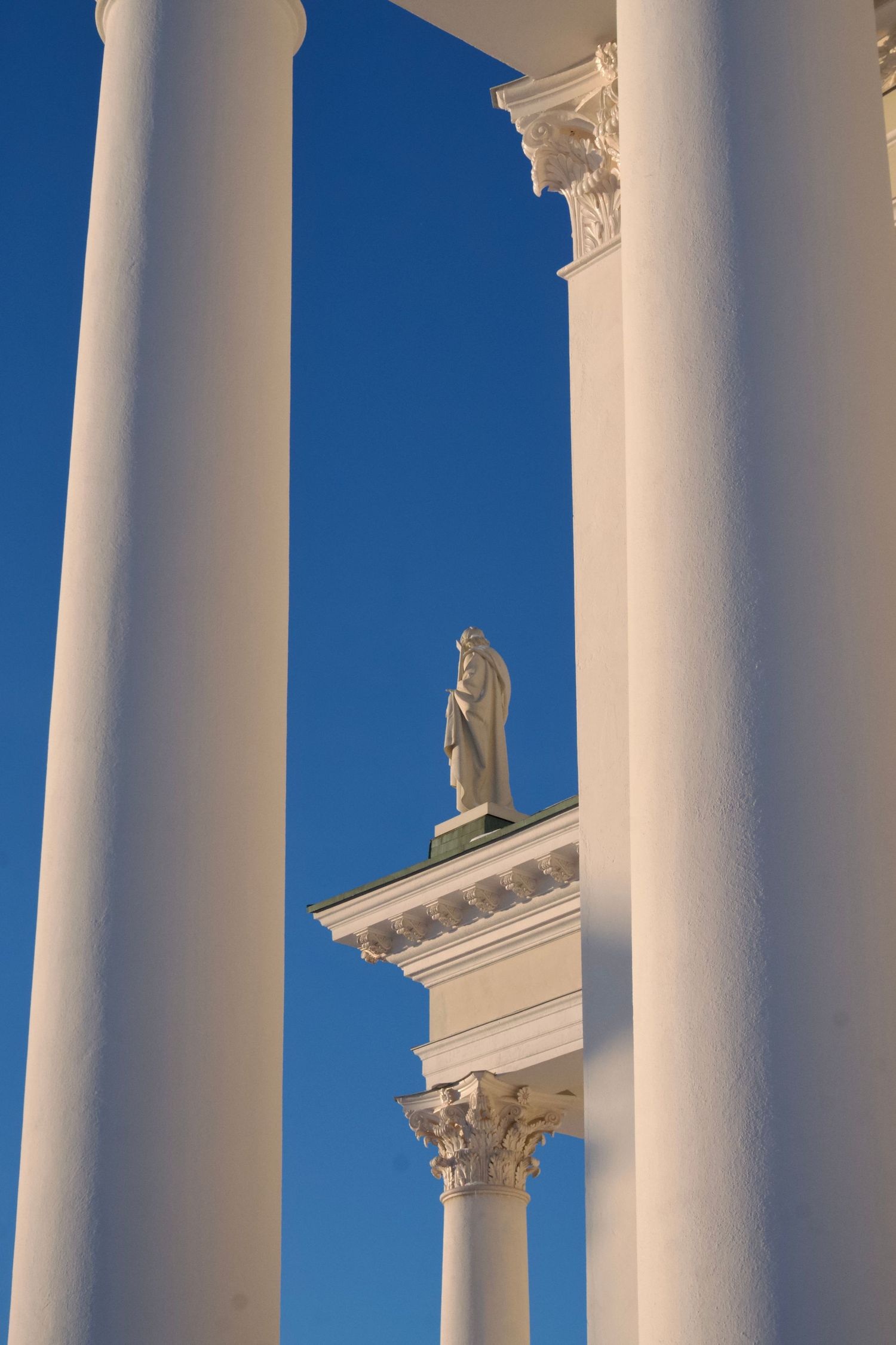White neoclassical columns frame a statue atop a cornice against a clear blue sky.
Reizen - Lynn - Helsinki