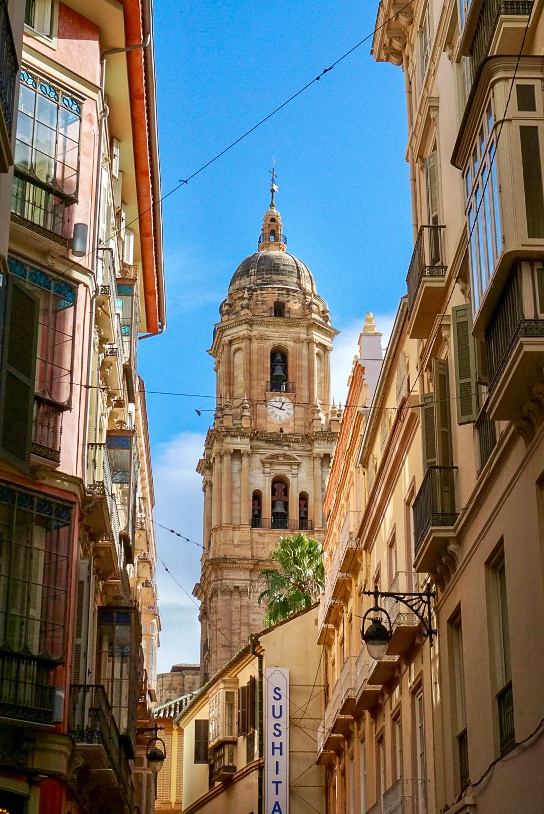 A narrow street in Málaga, Spain, framed by colorful buildings leading to the iconic Cathedral tower under a blue sky.
Lynn - Malaga