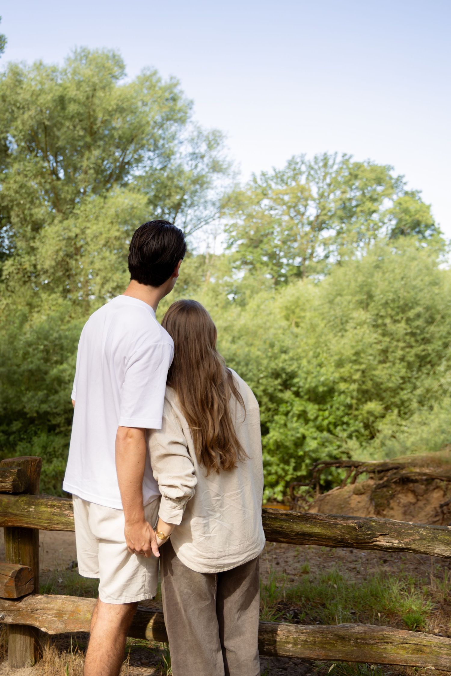 Couple holding hands while standing at a wooden fence, admiring a lush green forest landscape on a sunny day.
Indy en Daan - Koppelshoot
