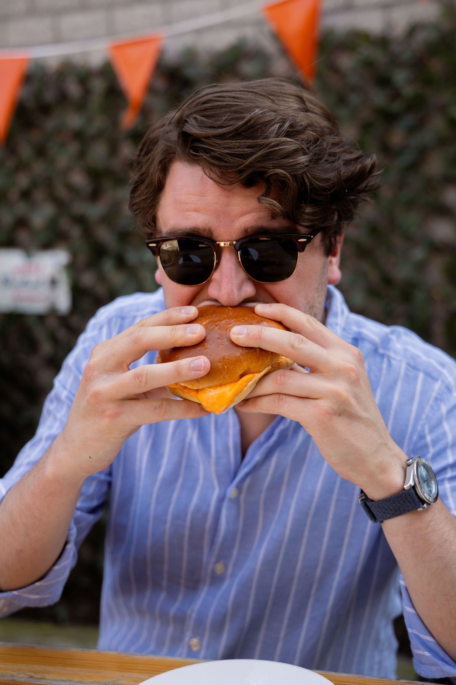 Man in sunglasses and blue striped shirt eating a burger outdoors at a table with orange bunting in background.
Yips - BBQ - Brandingshoot