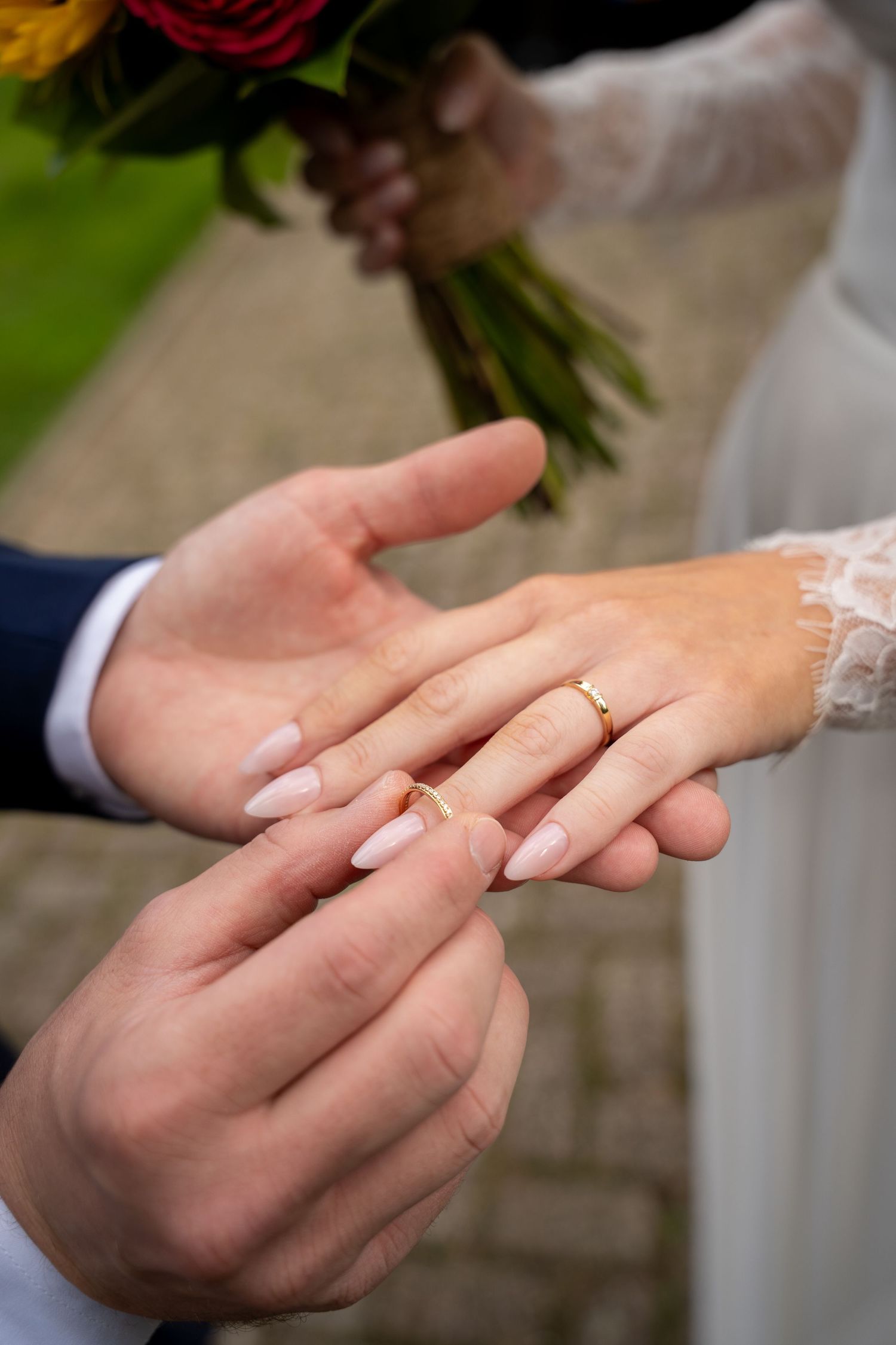 Groom placing a gold wedding ring on bride's finger, with bridal bouquet visible in background.