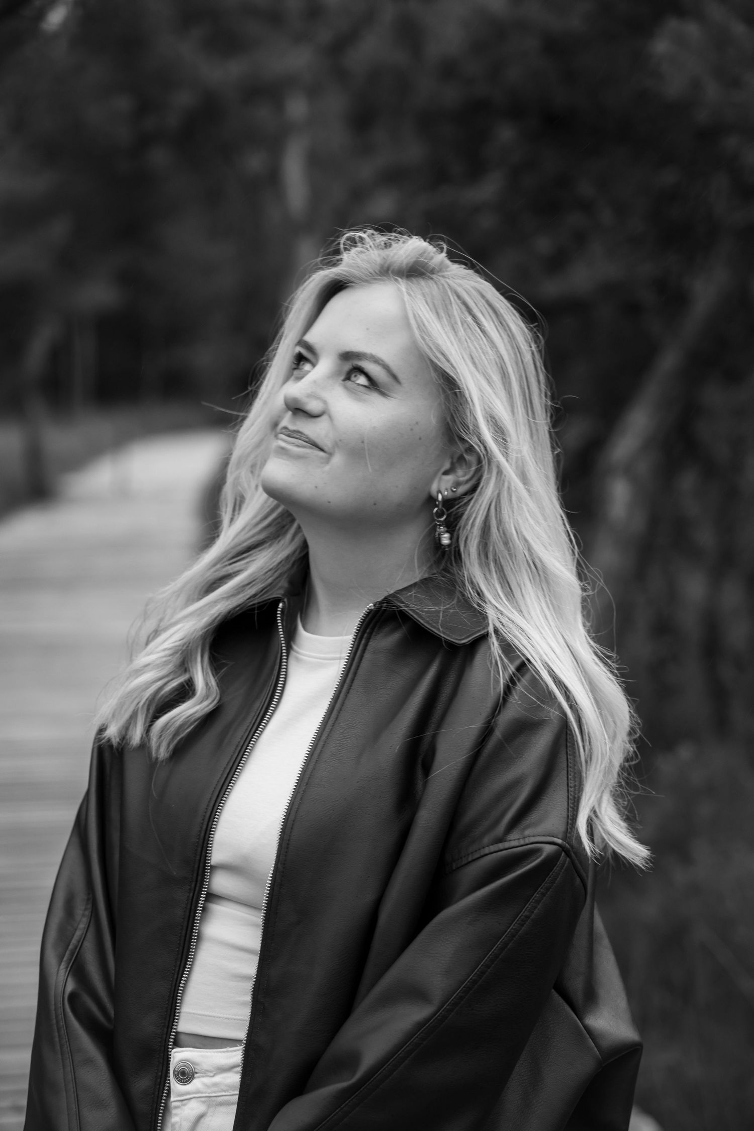 Black and white portrait of a blonde woman in a leather jacket smiling outdoors on a wooden path.