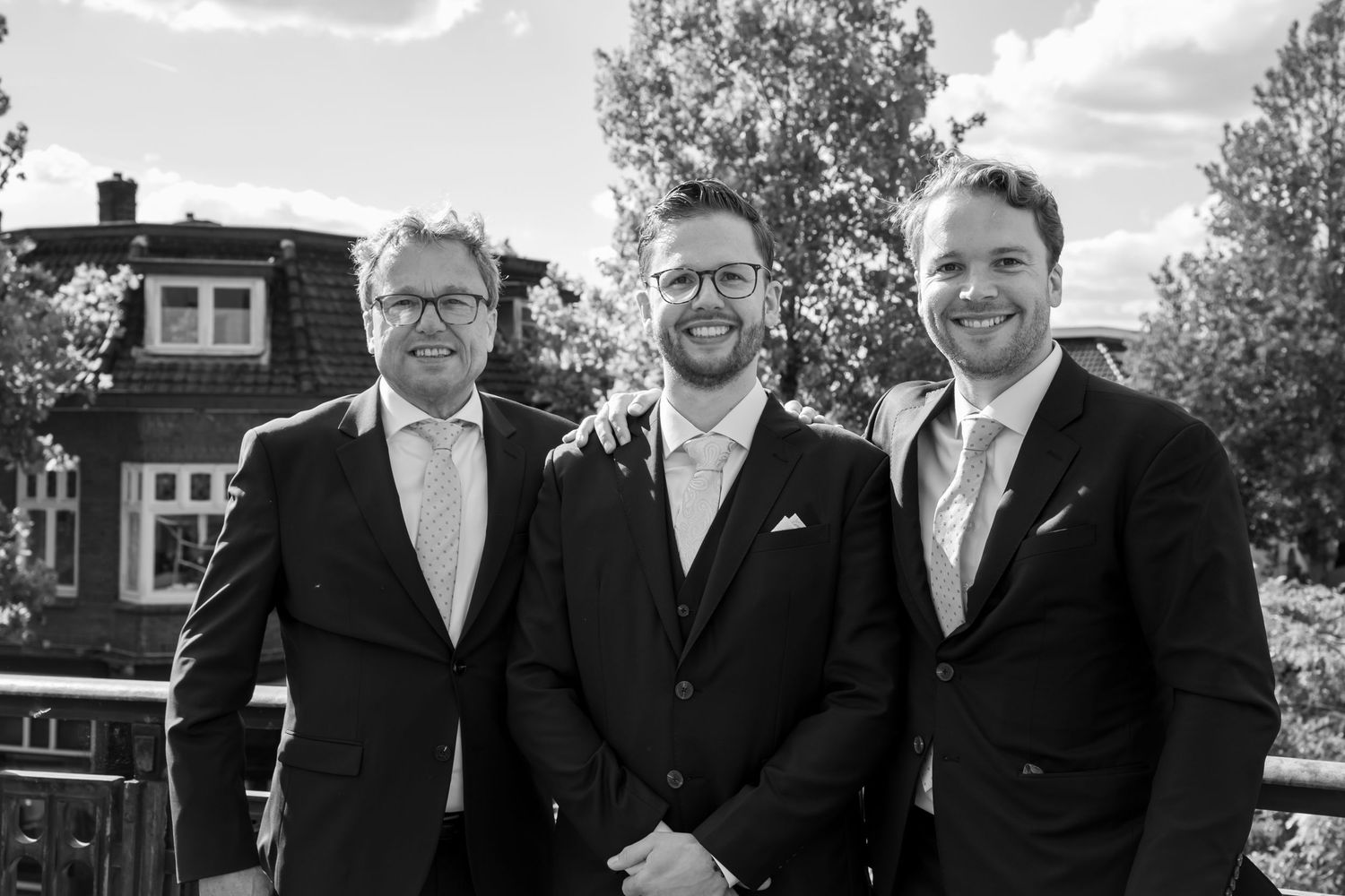 Three men in dark suits smiling outdoors in a black and white formal portrait photo.