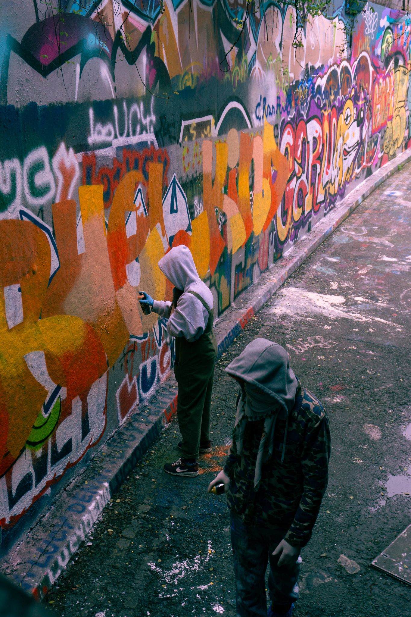 Two hooded graffiti artists spray painting a vibrant, colorful mural on a wall in an urban alleyway.
Reizen - Lynn - Londen