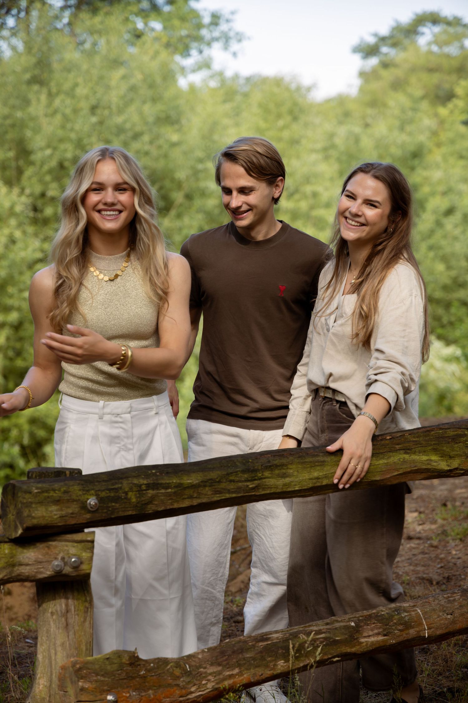 Three smiling young adults posing together on a wooden fence bridge surrounded by lush green trees outdoors.
Familie Wormgoor - Familieshoot