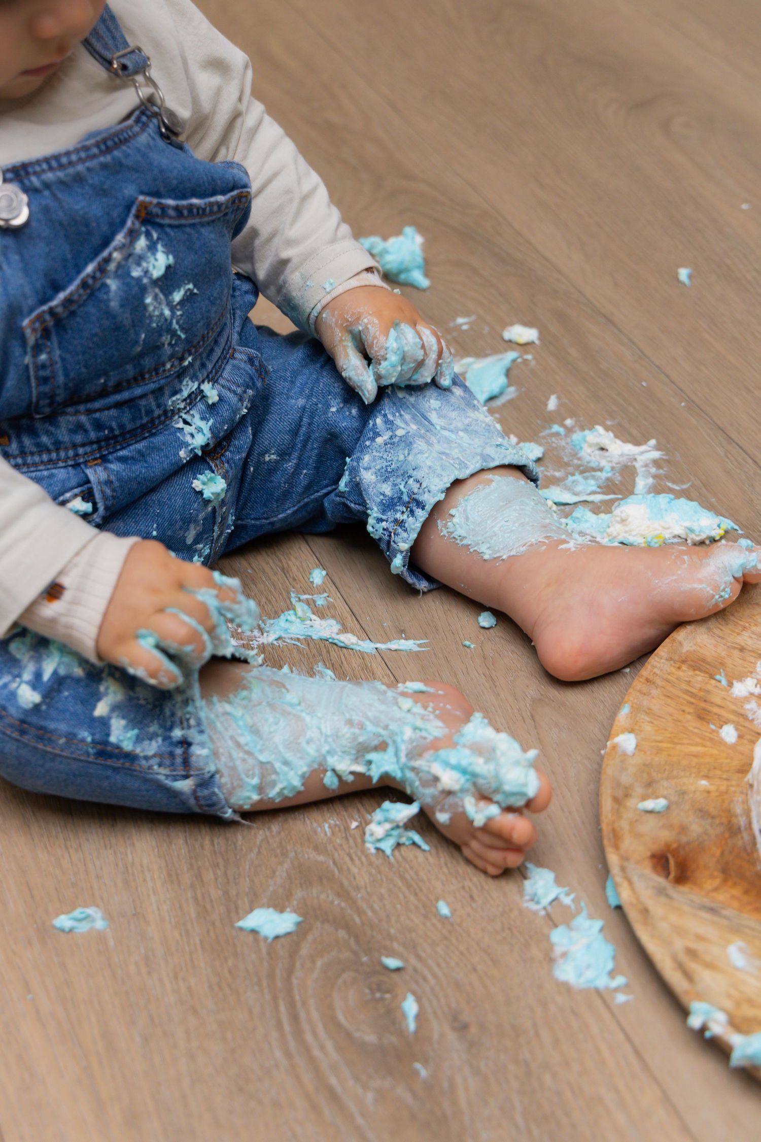 Toddler in denim overalls sitting on hardwood floor covered in smashed blue birthday cake.