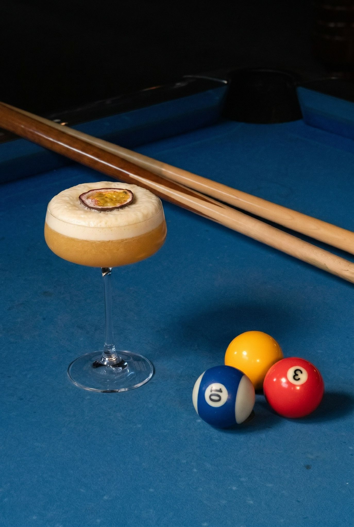 A frothy cocktail garnished with passion fruit sits on a blue pool table beside billiard balls and cue sticks.
Yip's Hengelo - Bowlen - Bran