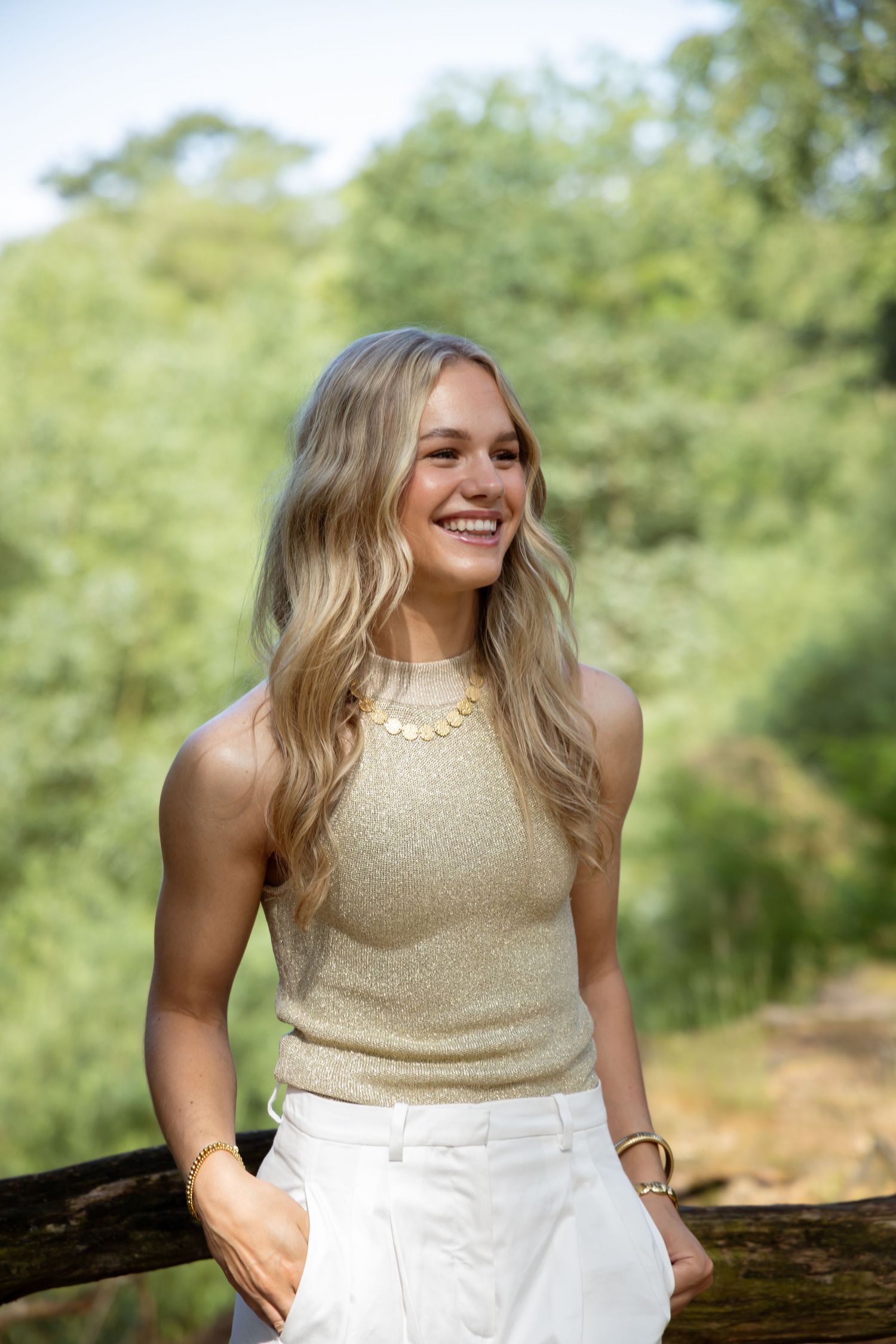Smiling blonde woman in gold tank top and white pants leaning on wooden fence outdoors.
Dewi - Solo foto