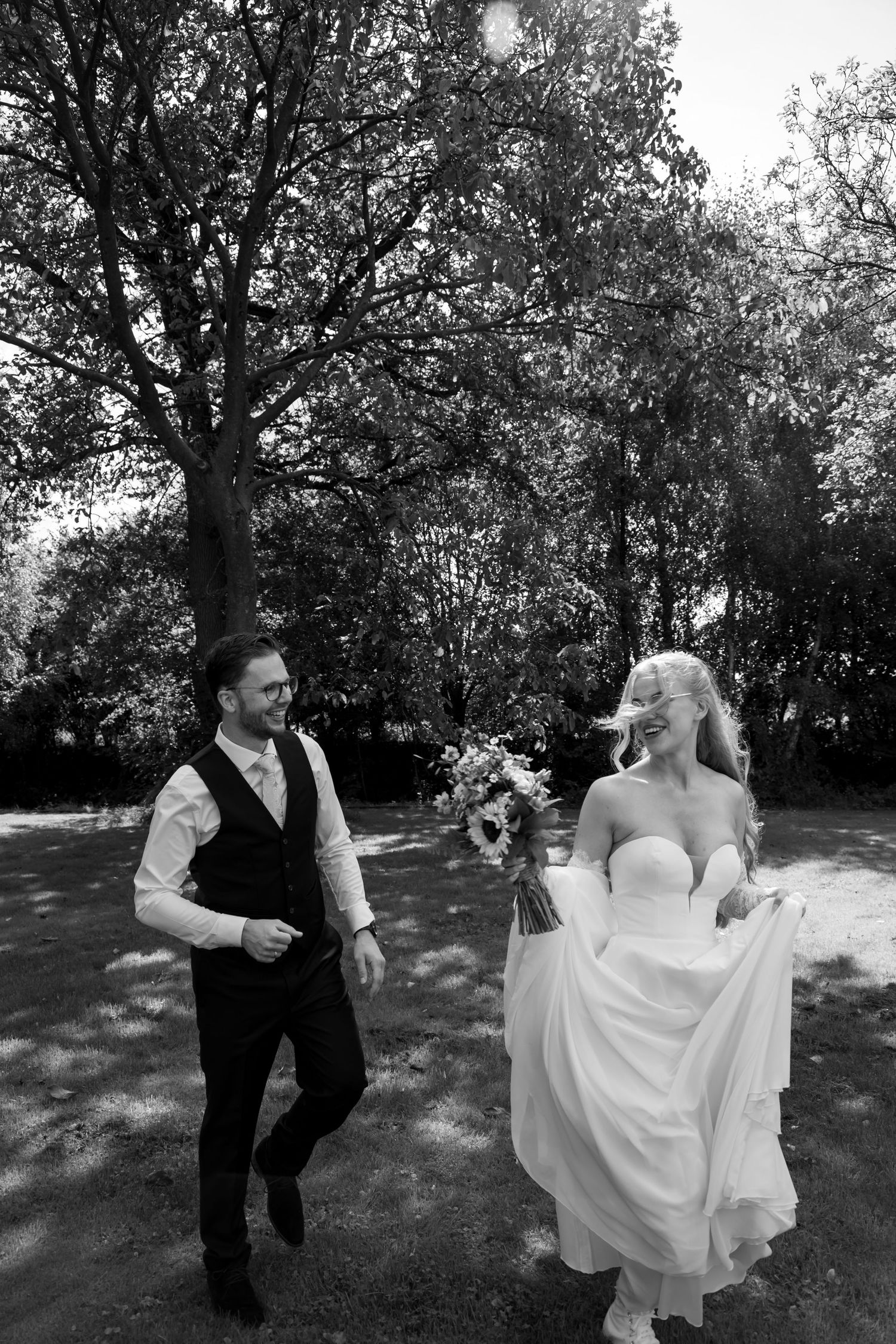 Black and white photo of a smiling bride in a strapless gown and groom in a vest walking together in a park.