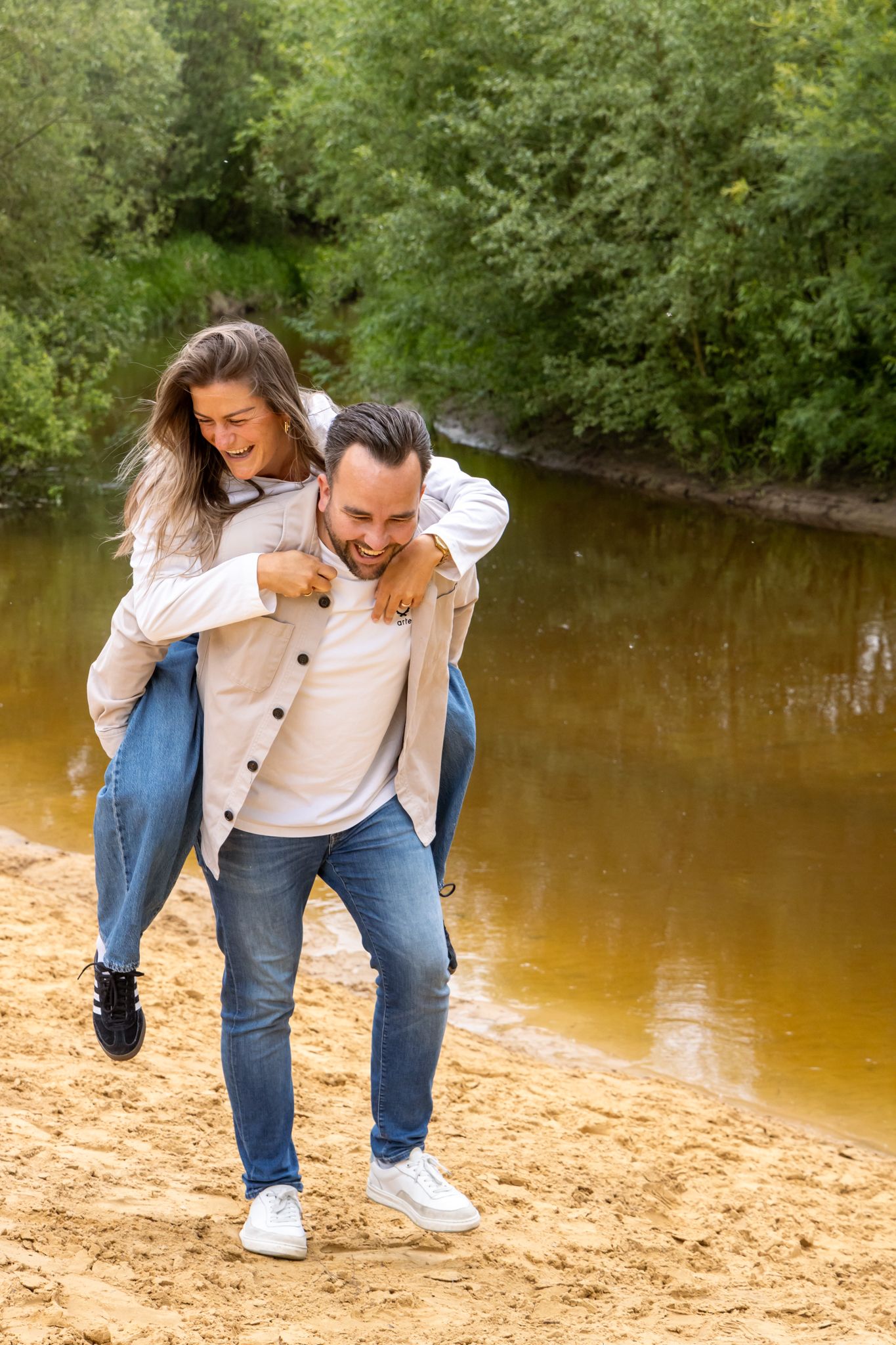 Happy couple enjoying a piggyback ride on a sandy riverbank surrounded by lush green trees.
Fleur en Timo - Koppelshoot