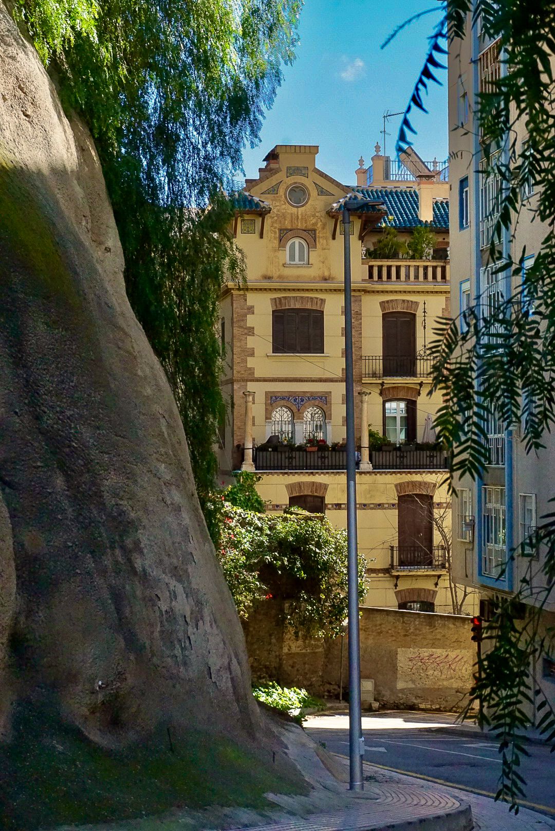Historic yellow European building with ornate balconies framed by lush trees and a large rock in a sunny courtyard.
Reizen - Lynn - Malaga