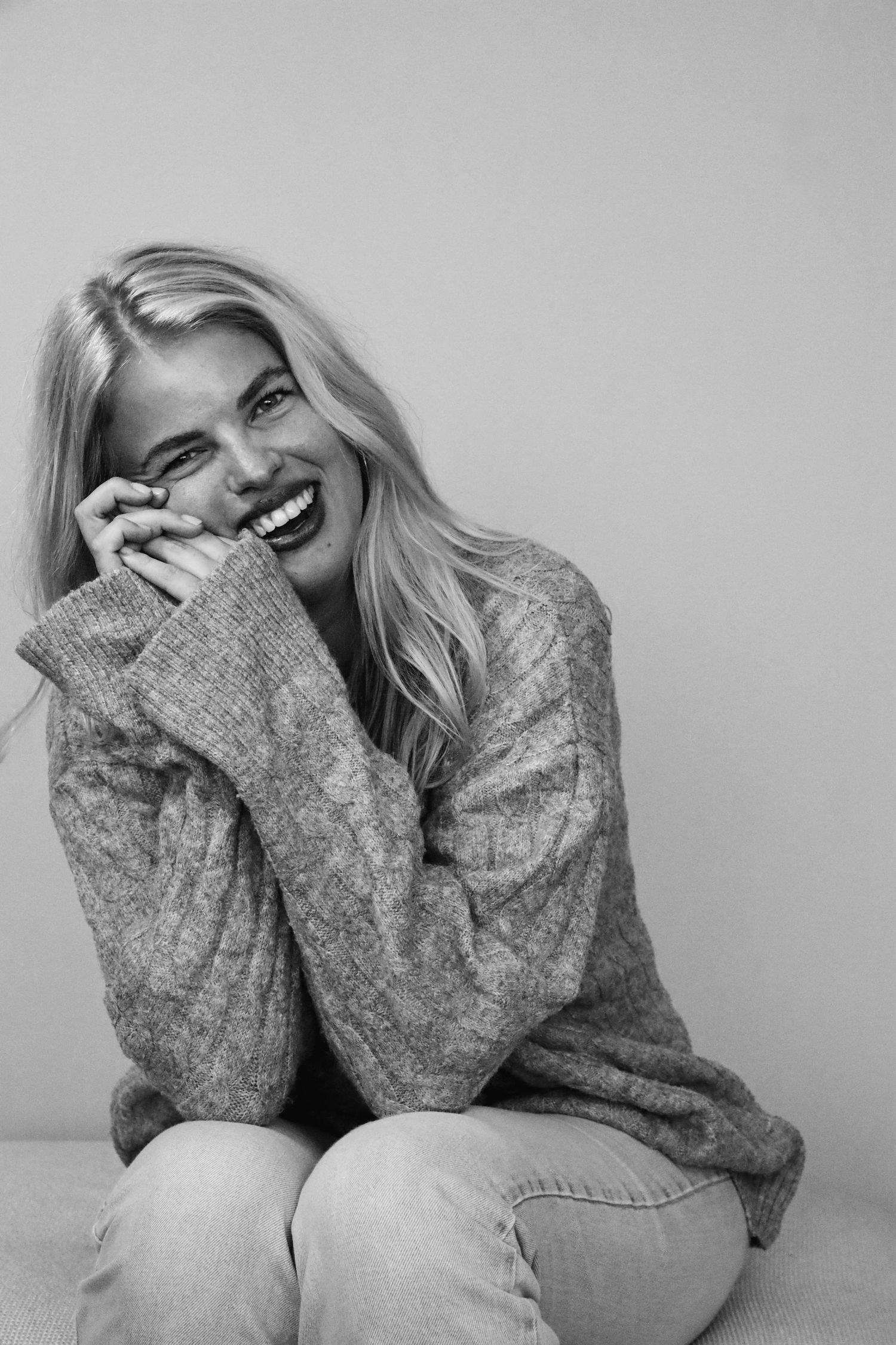 Laughing blonde woman in cozy knit sweater and jeans, sitting relaxed in black and white portrait photo.
Tess - Soloshoot