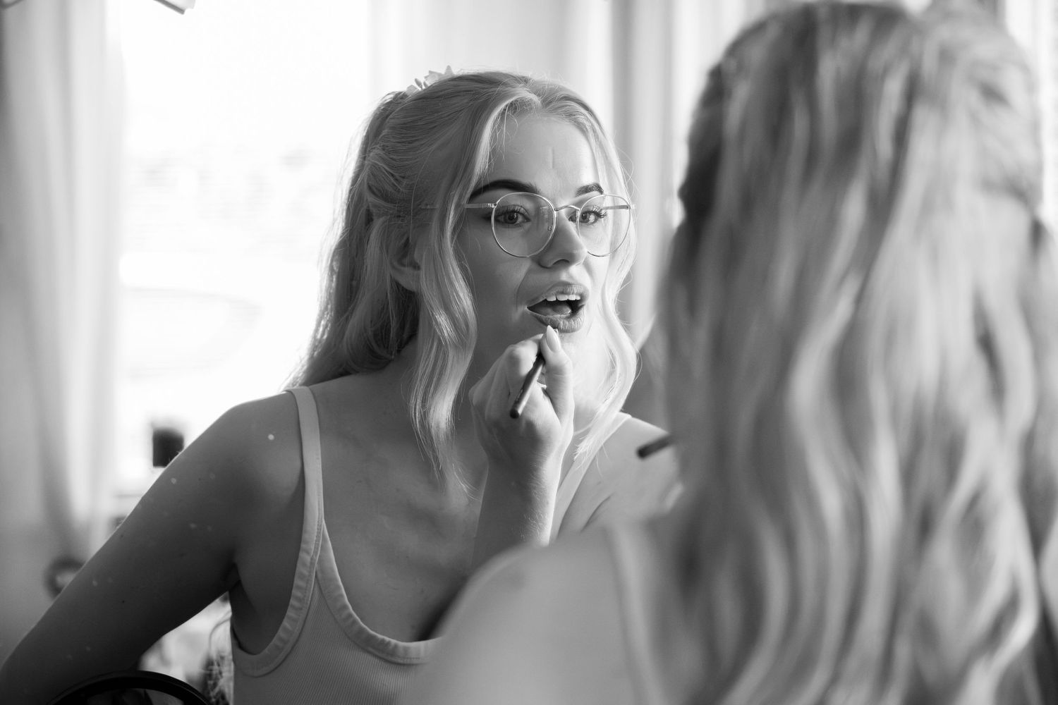 Black and white photo of a young woman with glasses applying her lipstick.