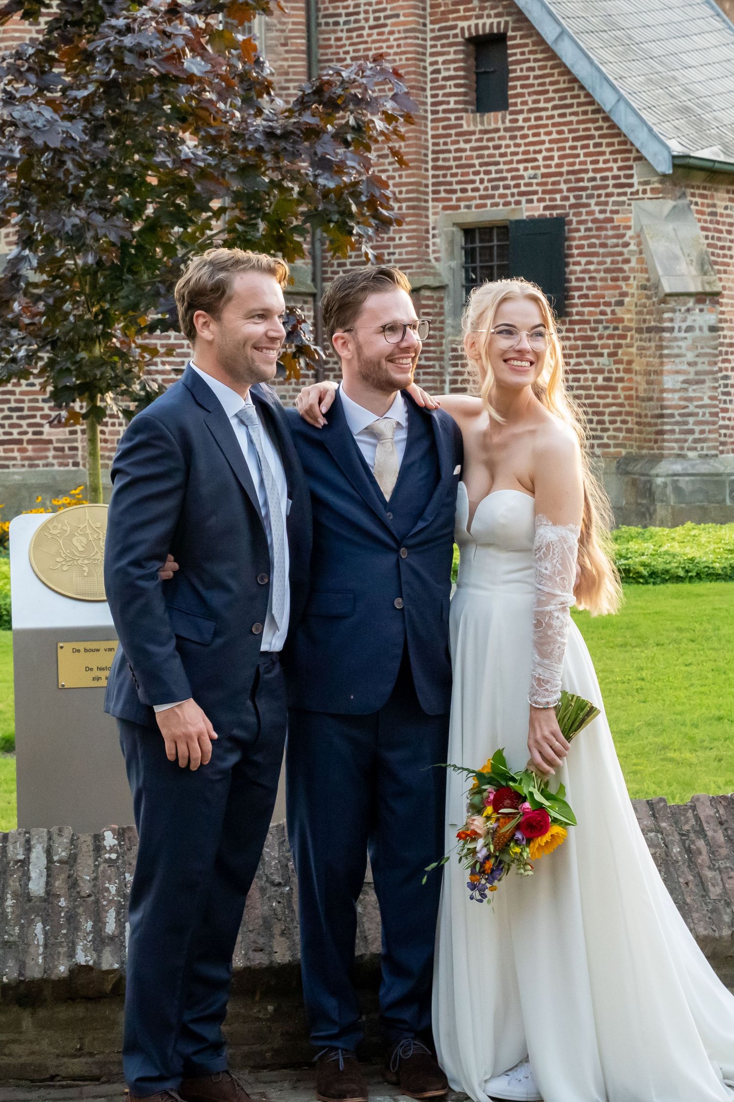 Bride in white gown with bouquet poses with two men in navy suits at a brick building wedding venue.