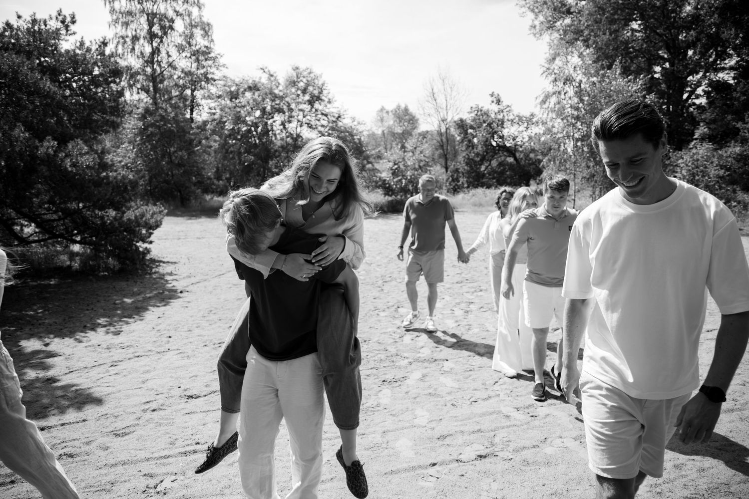 Black and white photo of family enjoying a sunny outdoor gathering, with a woman having a piggyback ride on a sandy path.
Familie Wormgoor